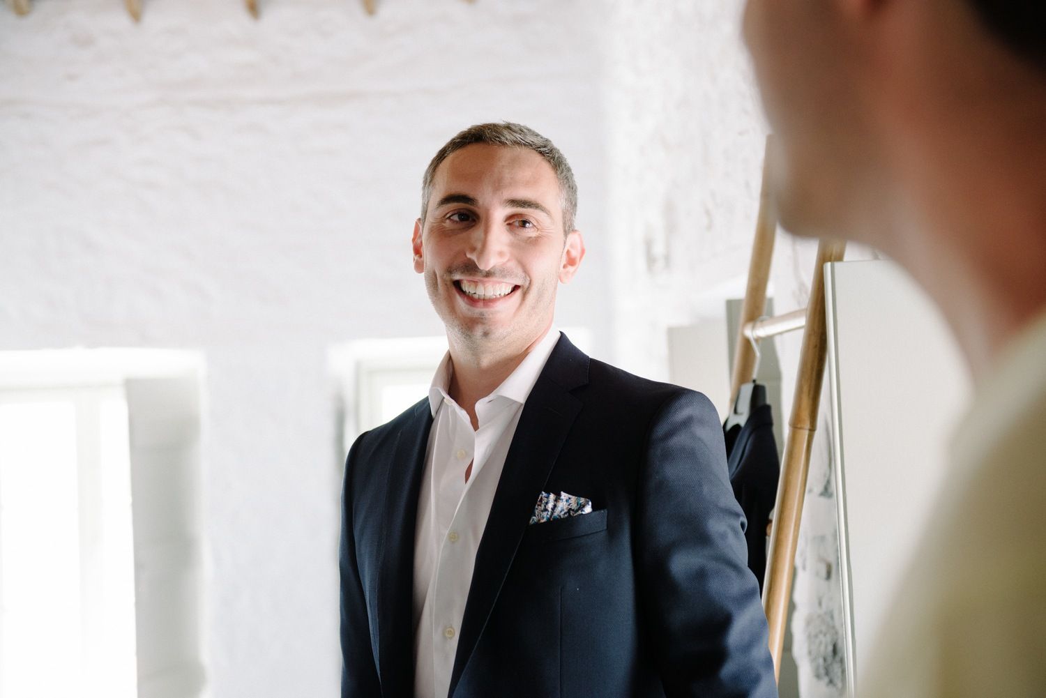 A man in a navy blazer smiles warmly in a bright interior setting.