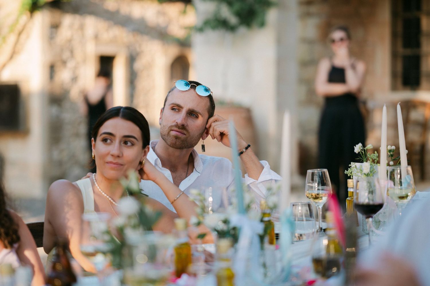 Two people seated at an outdoor dinner table with wine glasses and decorative blue floral centerpieces.