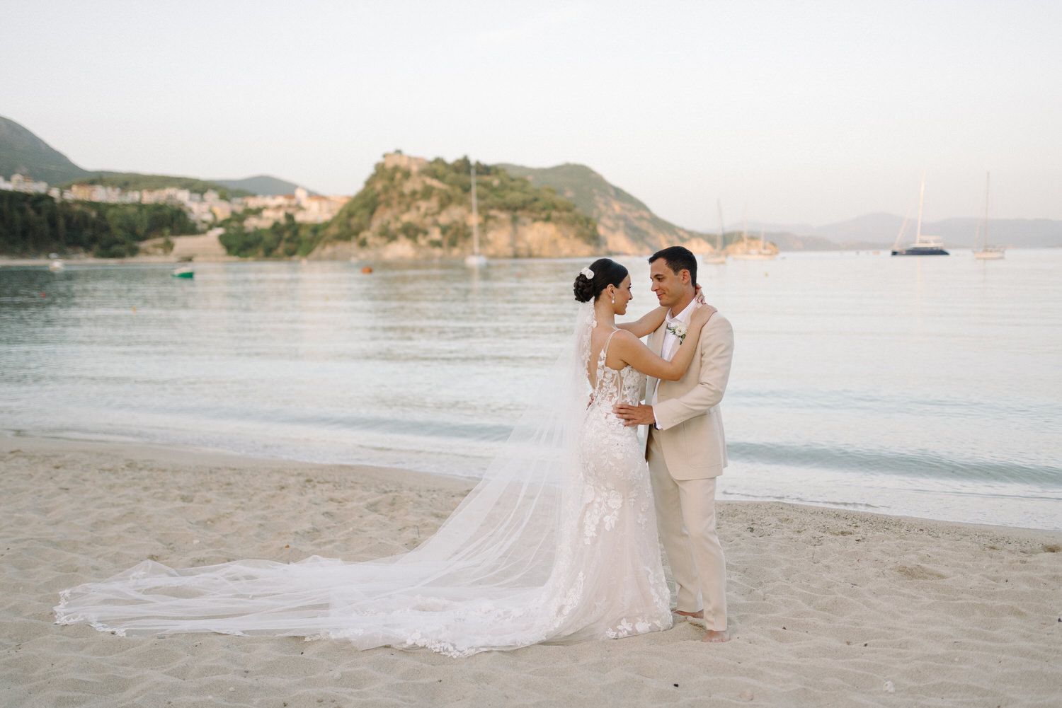 A romantic beach wedding photo with a couple embracing against a scenic coastal backdrop with mountains and water.