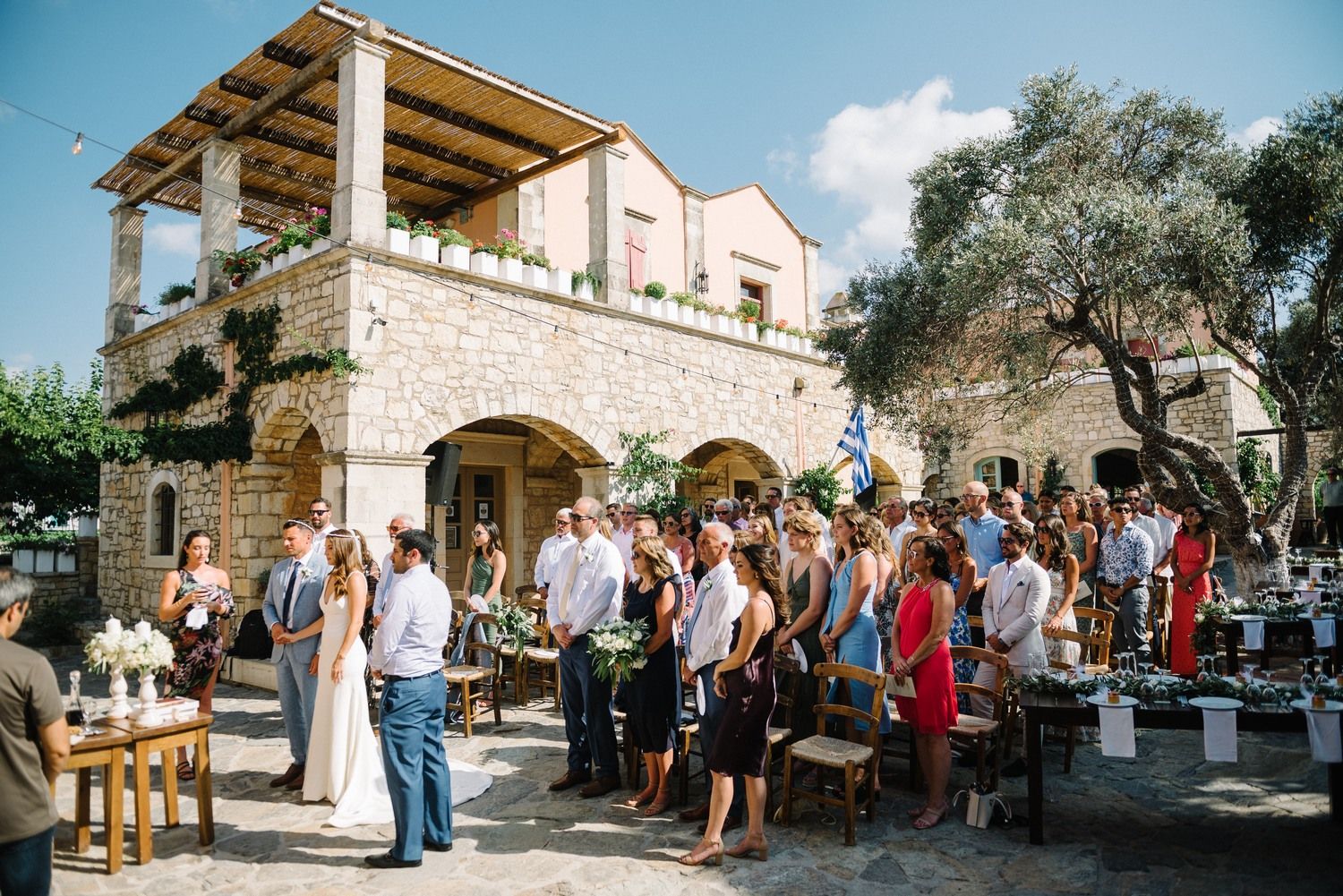 Outdoor wedding reception gathering at a rustic stone villa with guests mingling on a sunny day.