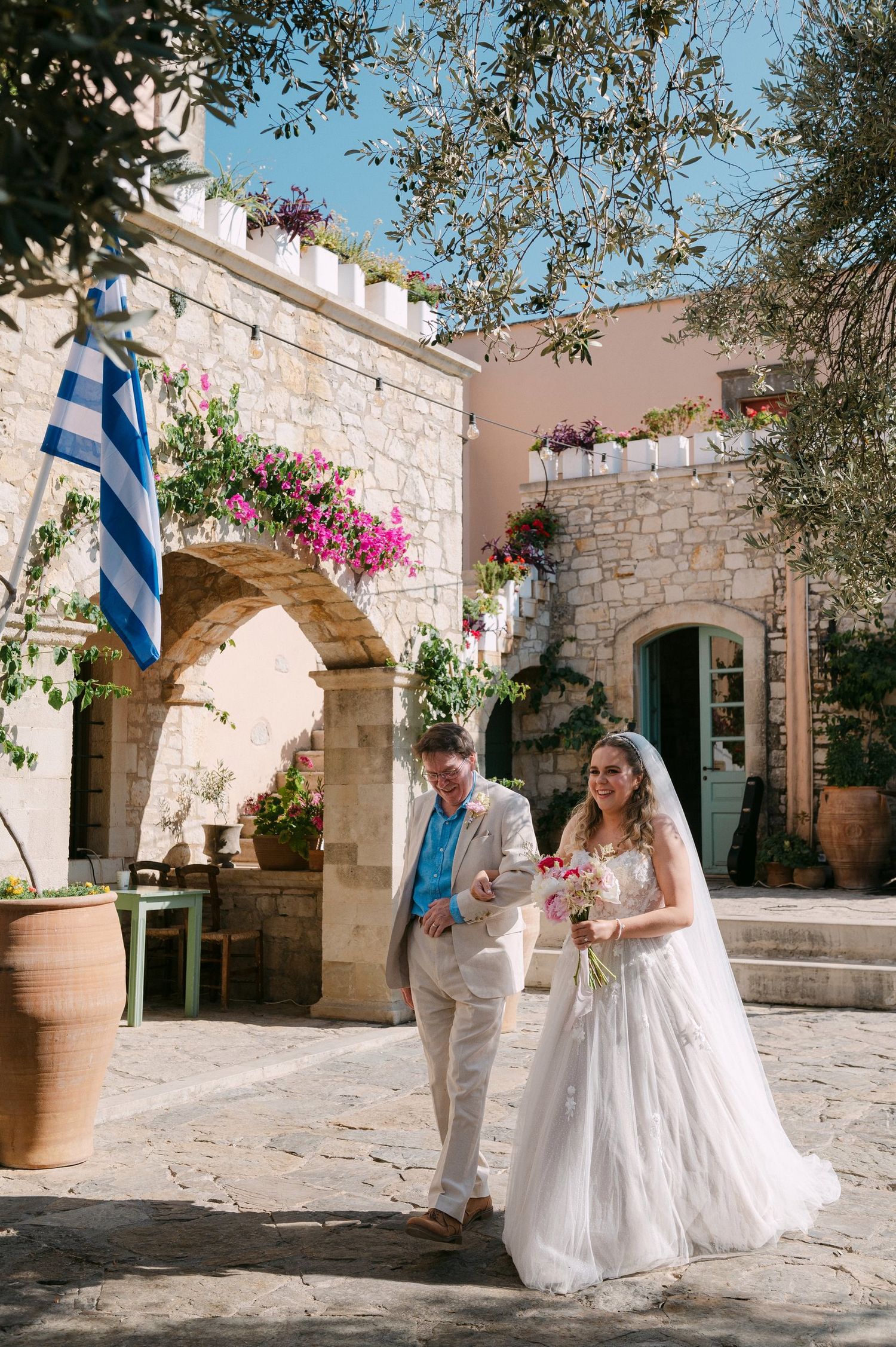 A wedding celebration at a traditional stone building in Greece with a Greek flag and pink bougainvillea flowers.