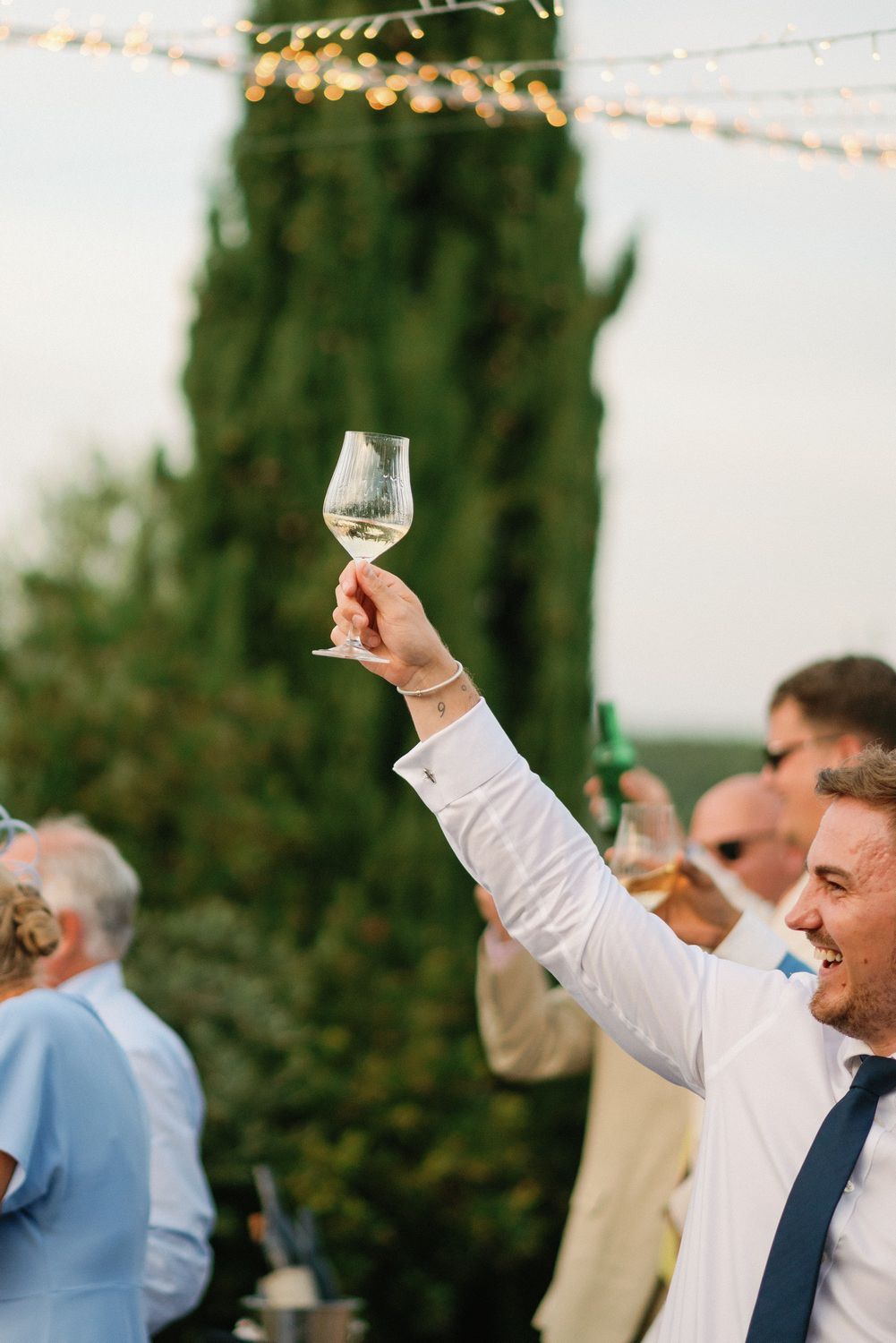 Guest raises a wine glass in celebration during an outdoor wedding reception with string lights.