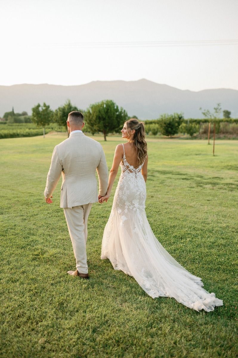 A couple walks across a green lawn with mountains in background, bride's lace dress trailing behind.