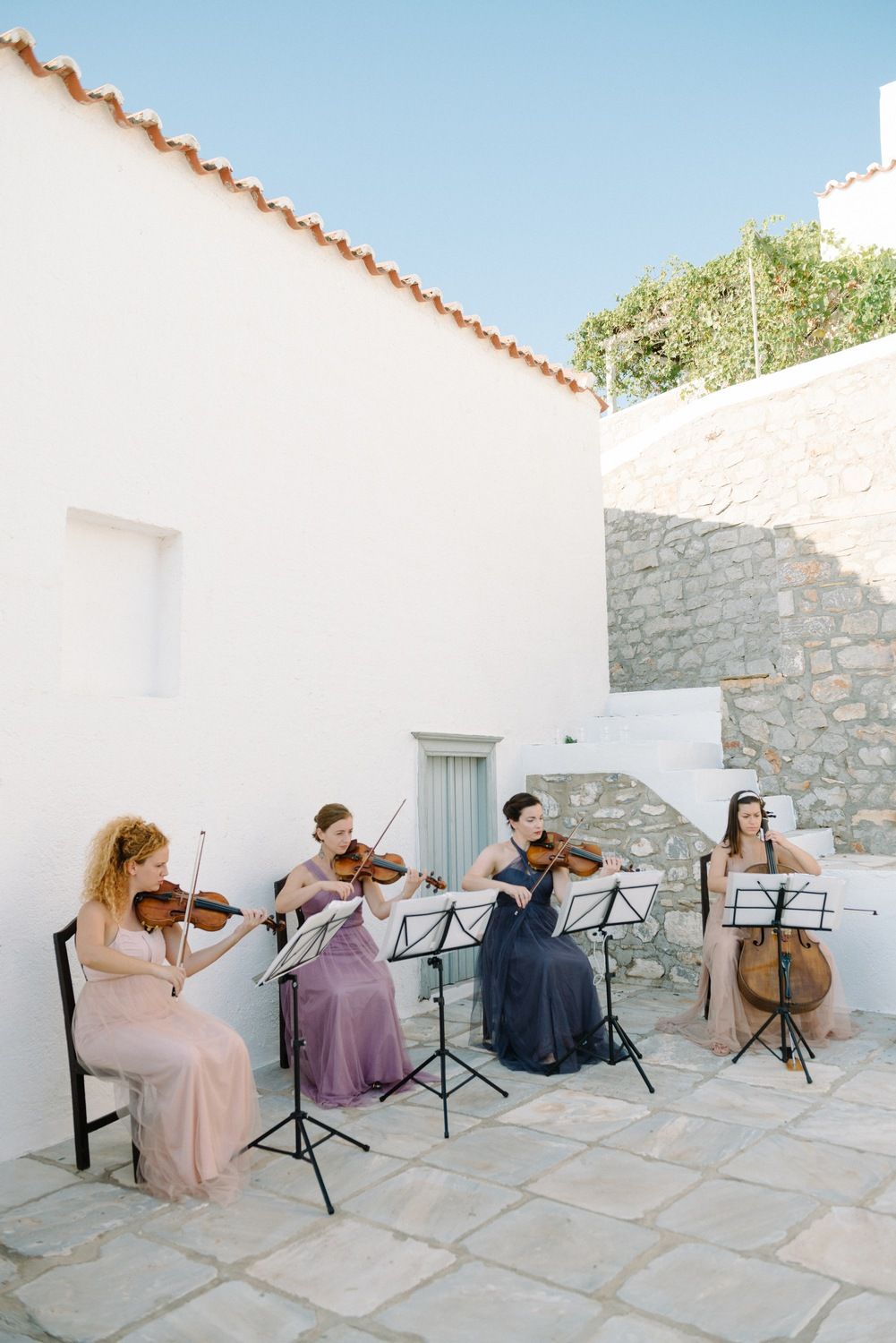 String quartet performs in elegant dresses on a Mediterranean terrace with white walls and stone flooring.