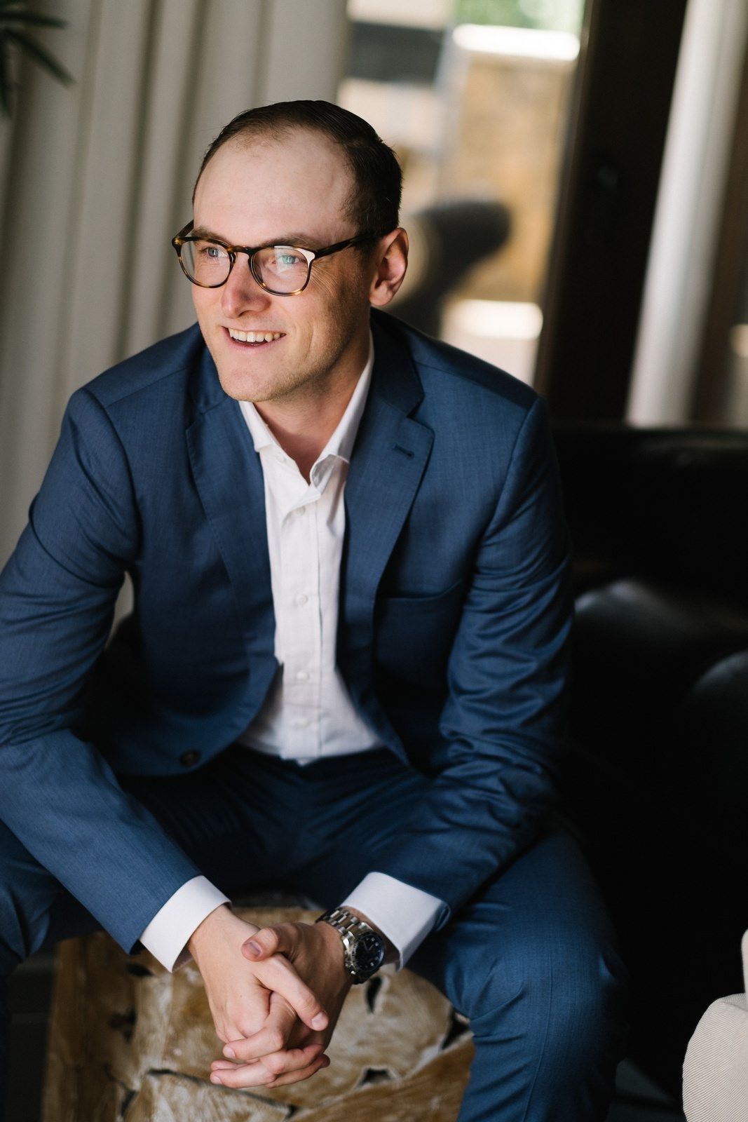 A person in a navy suit and glasses sits smiling on a couch.