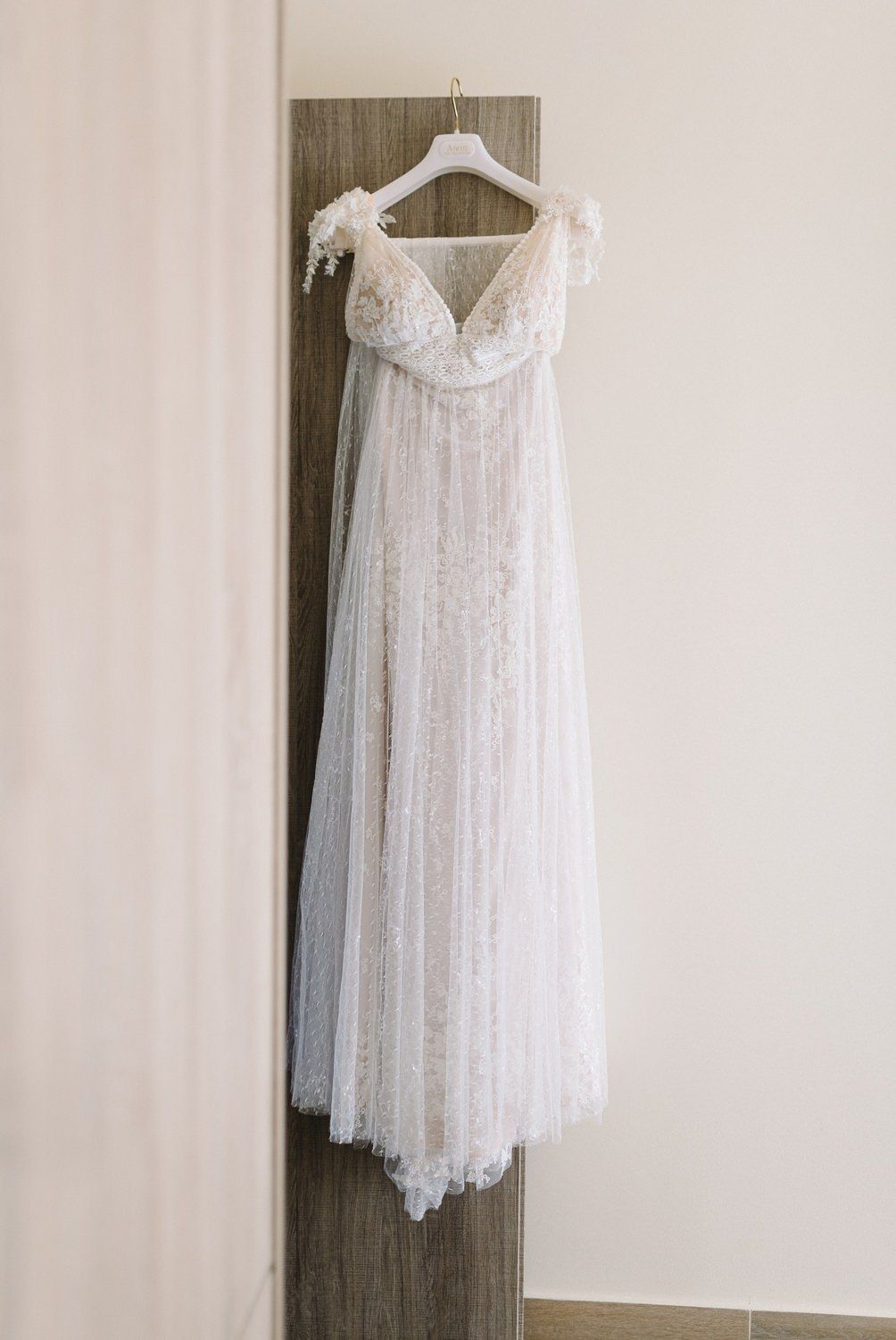 Elegant white lace wedding dress with empire waist and delicate tulle details hanging on wooden wall.