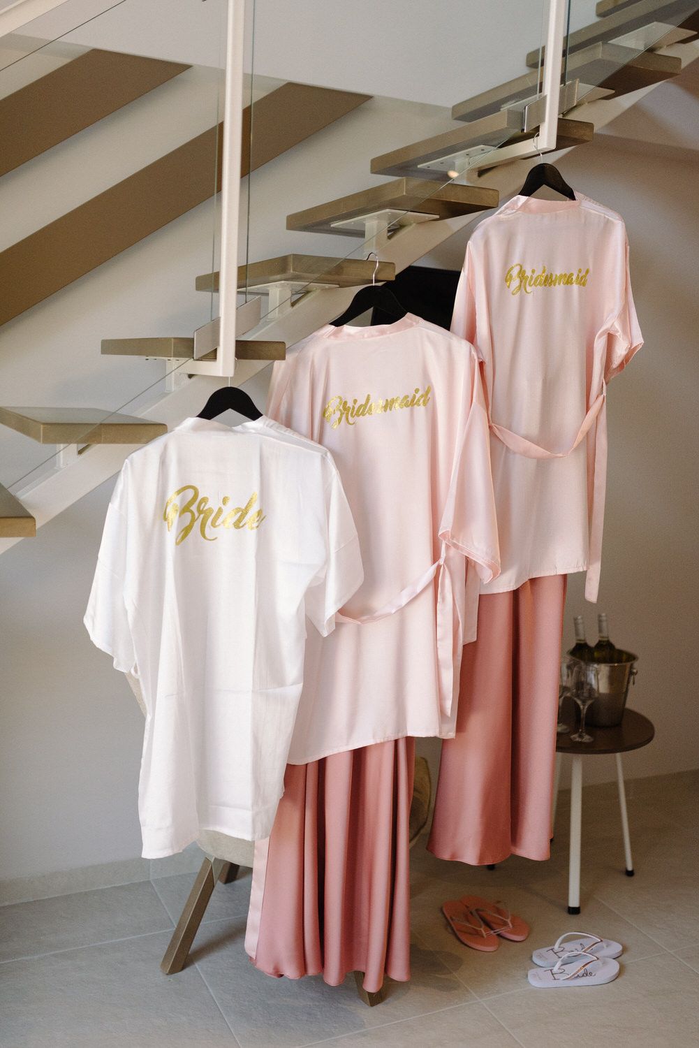 Wedding party robes in white and pink with gold lettering hanging on wooden staircase.