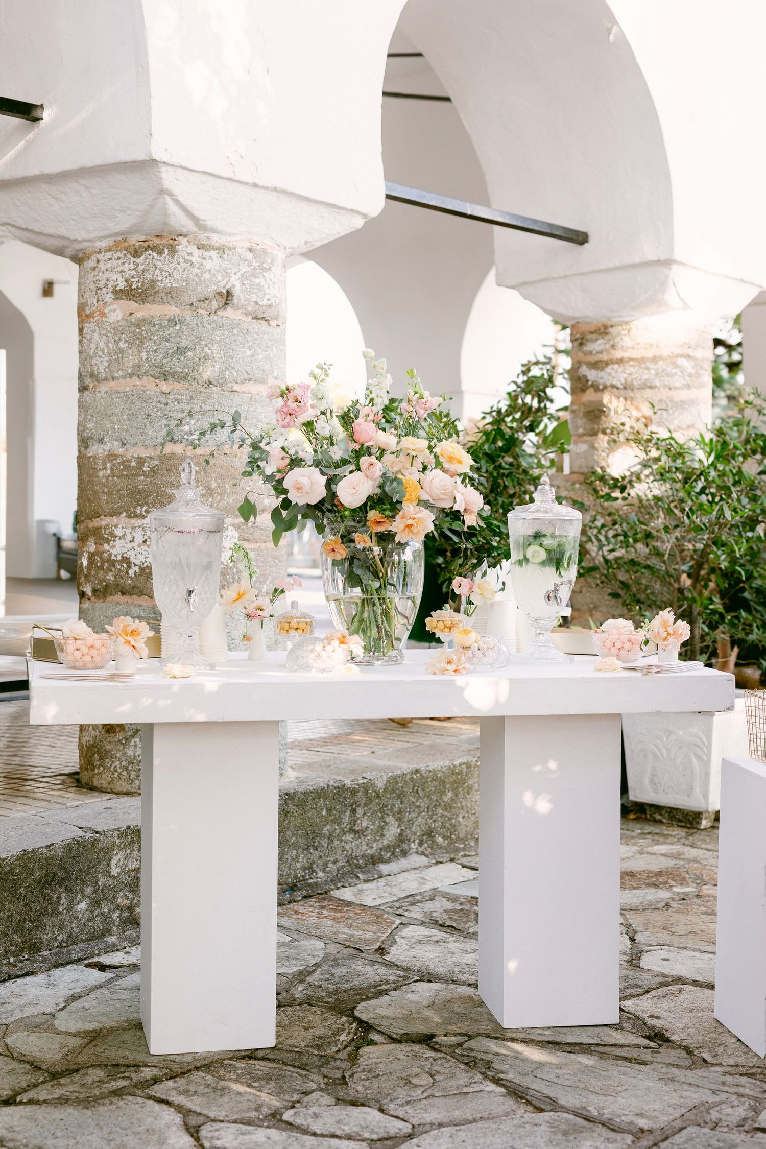 Elegant wedding dessert table with white tablecloth and pink floral arrangements under Mediterranean-style stone archways.