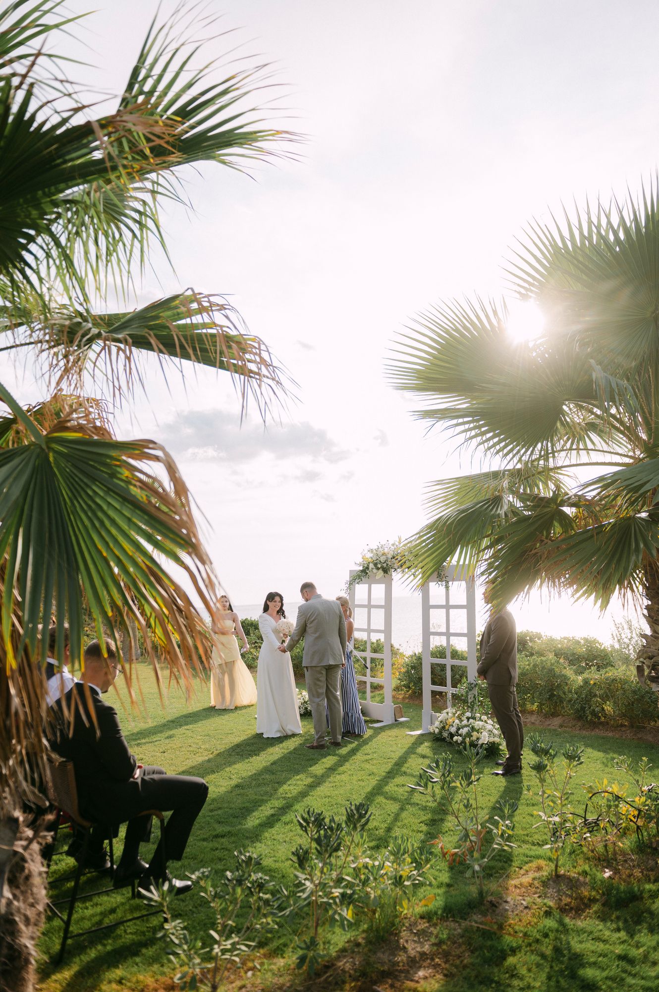 An outdoor wedding ceremony setup with palm trees and a white arch on a grassy hilltop during golden hour at Halkidiki Greeece.
