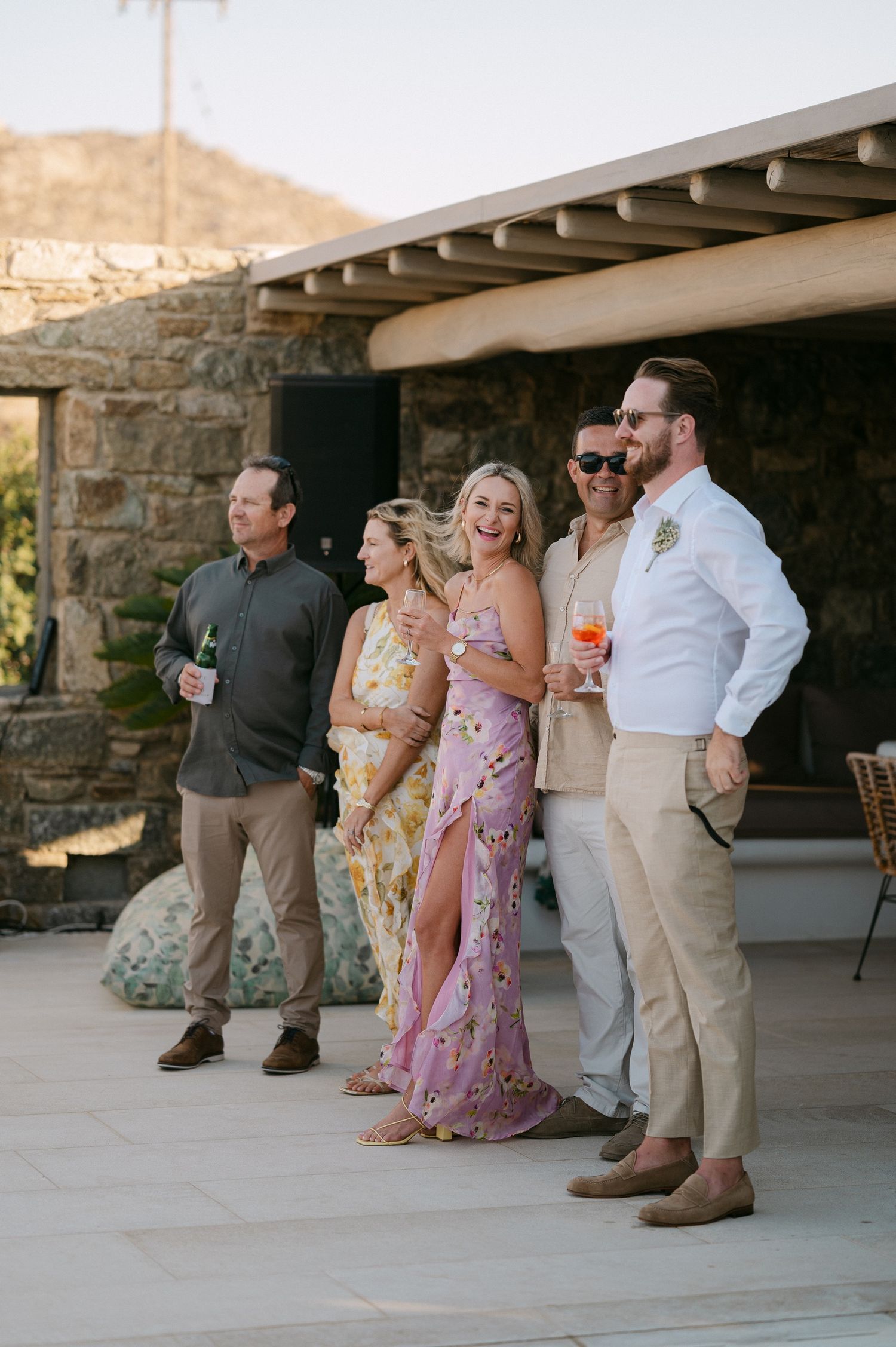 A group of wedding guests in casual attire gather on a covered stone patio during a relaxed outdoor reception.
