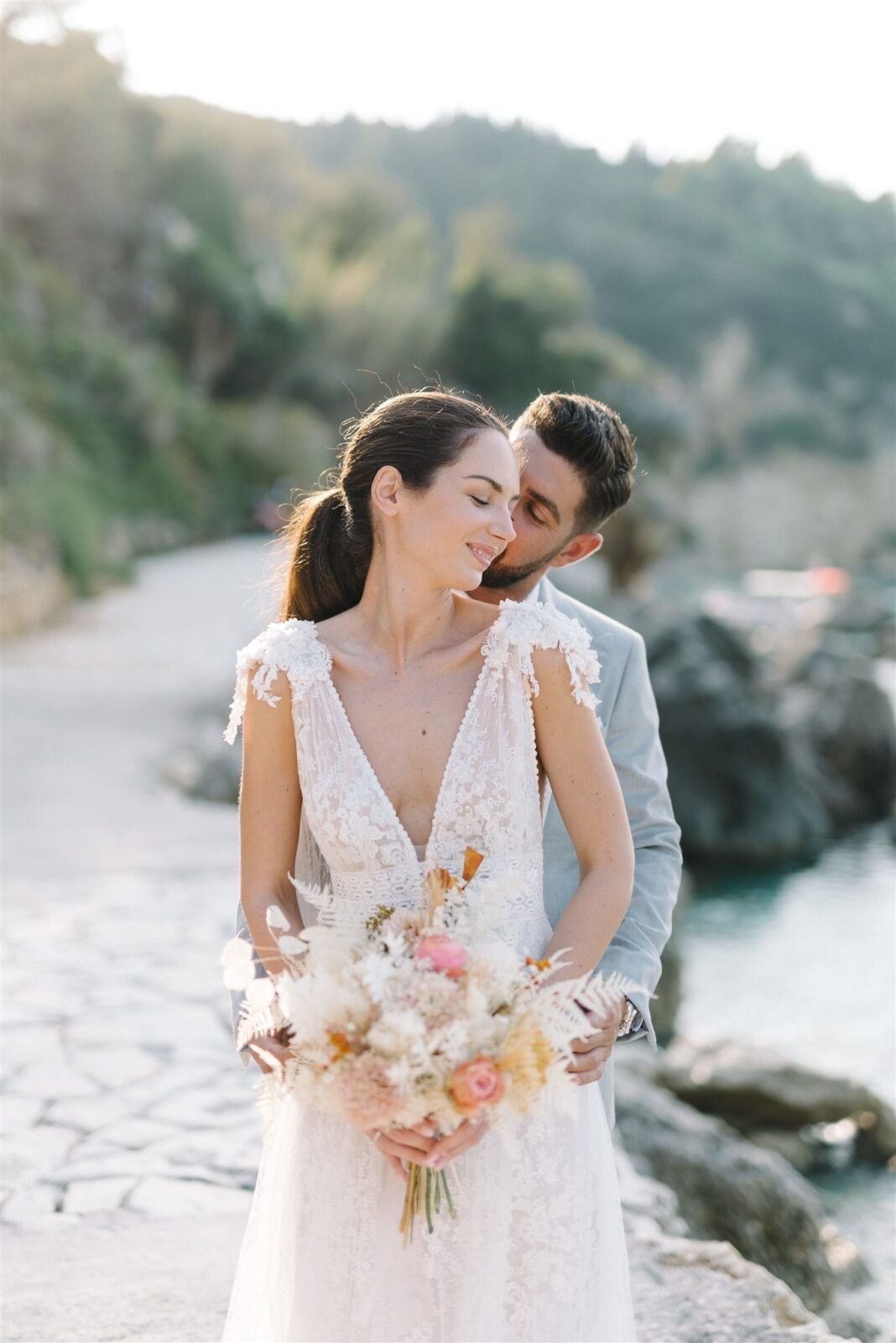 Romantic wedding couple embraces on a rocky beach with soft pastel bouquet at sunset.
