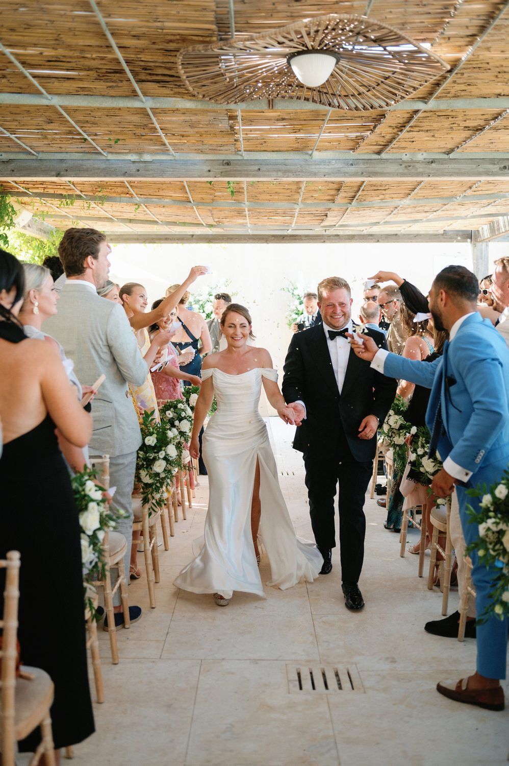 Wedding celebration in a rustic covered walkway decorated with floral arrangements and wooden chairs.