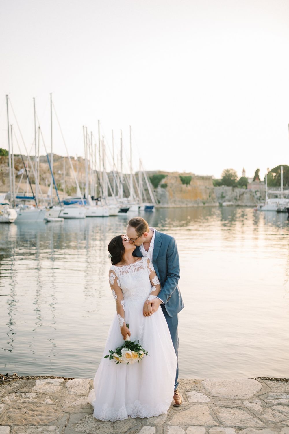 A romantic wedding couple embraces by a marina with sailboats and stone walls at sunset.
