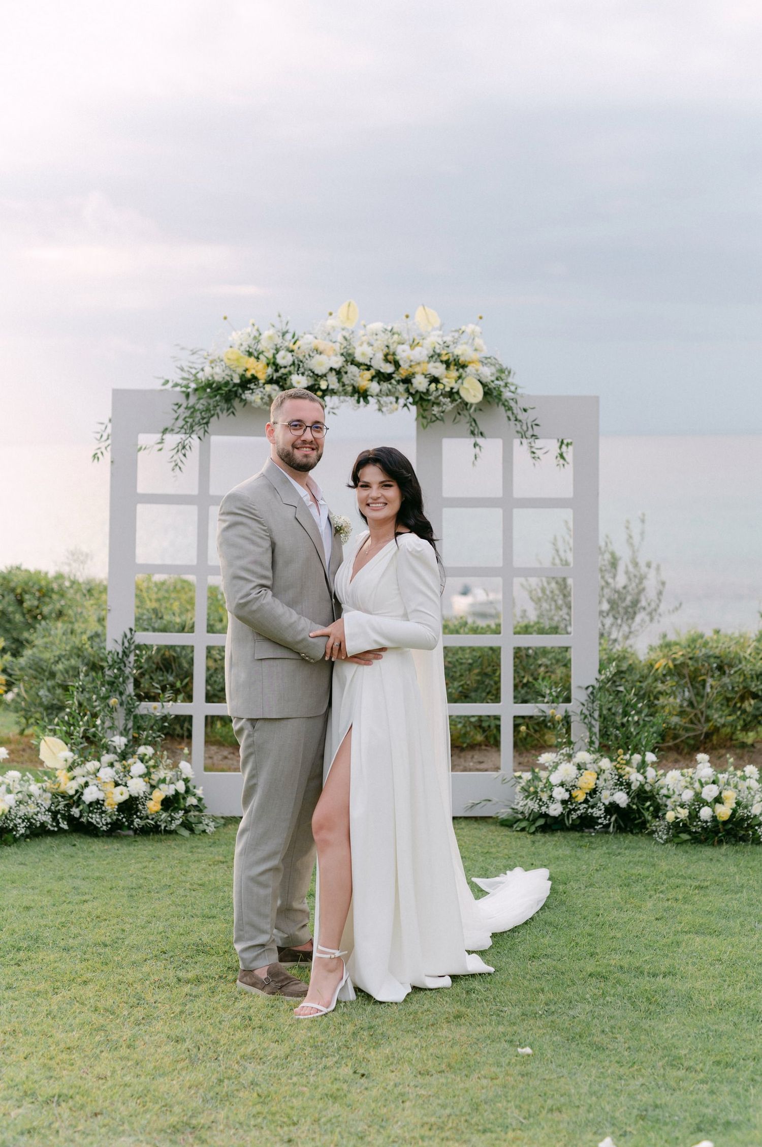 Couple poses together in front of white wedding arch decorated with floral arrangements on grassy outdoor venue.