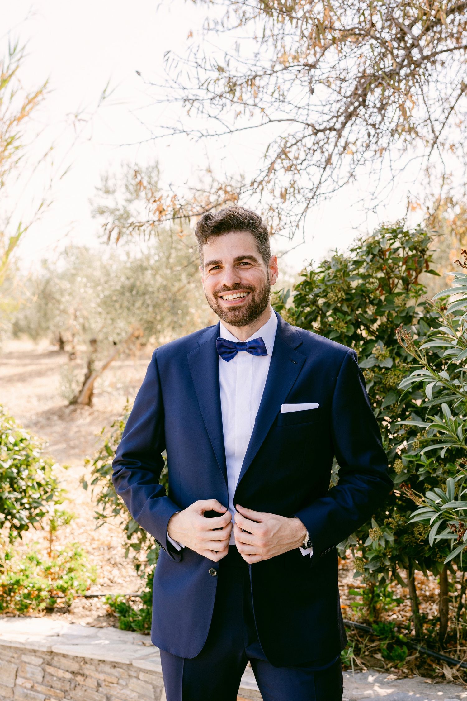 A groom in a navy blue tuxedo with bow tie stands smiling in an outdoor garden setting during golden hour.