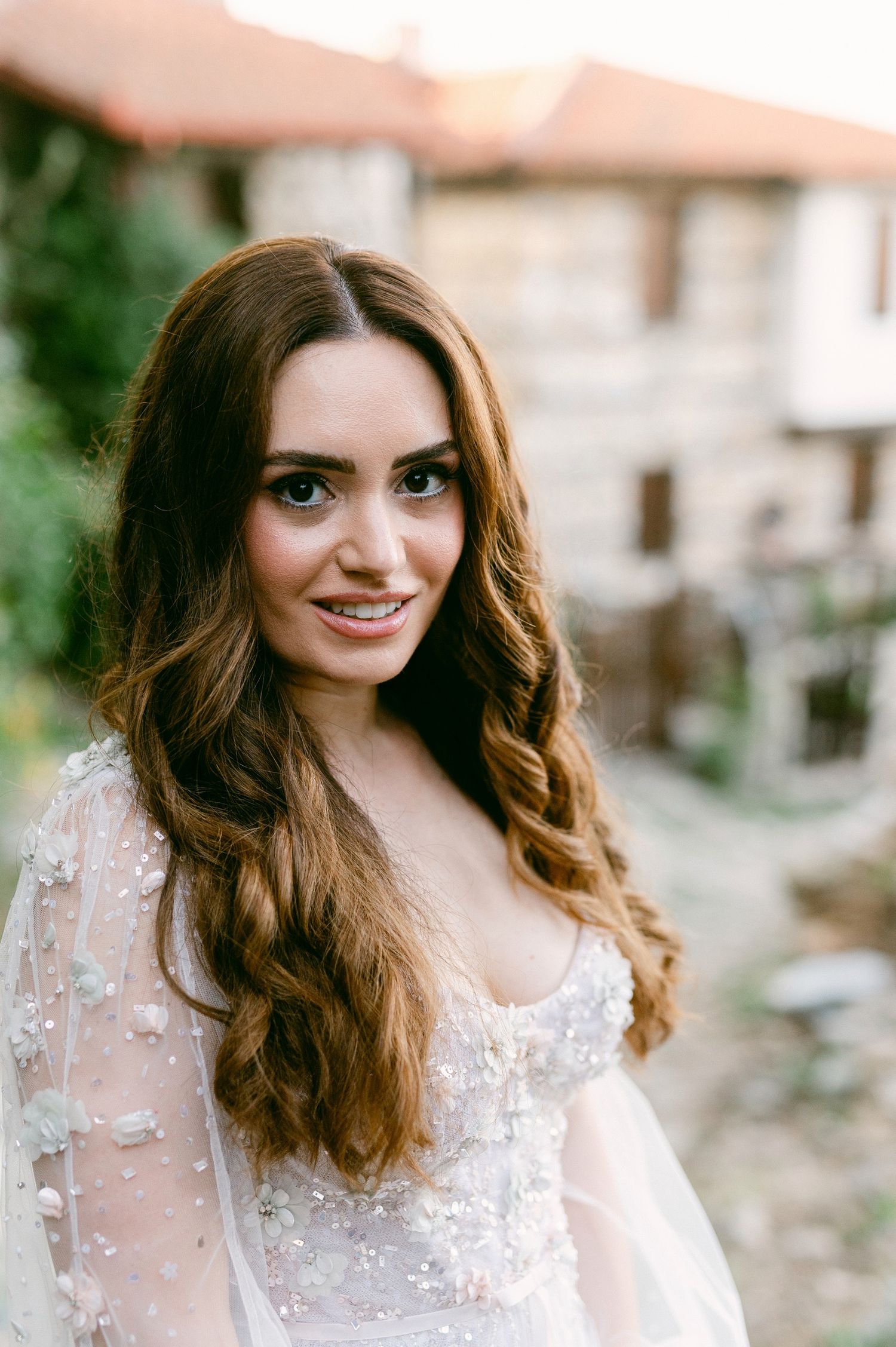 A woman in a delicate beaded white dress with sheer sleeves and long wavy brown hair smiles at the camera outdoors.