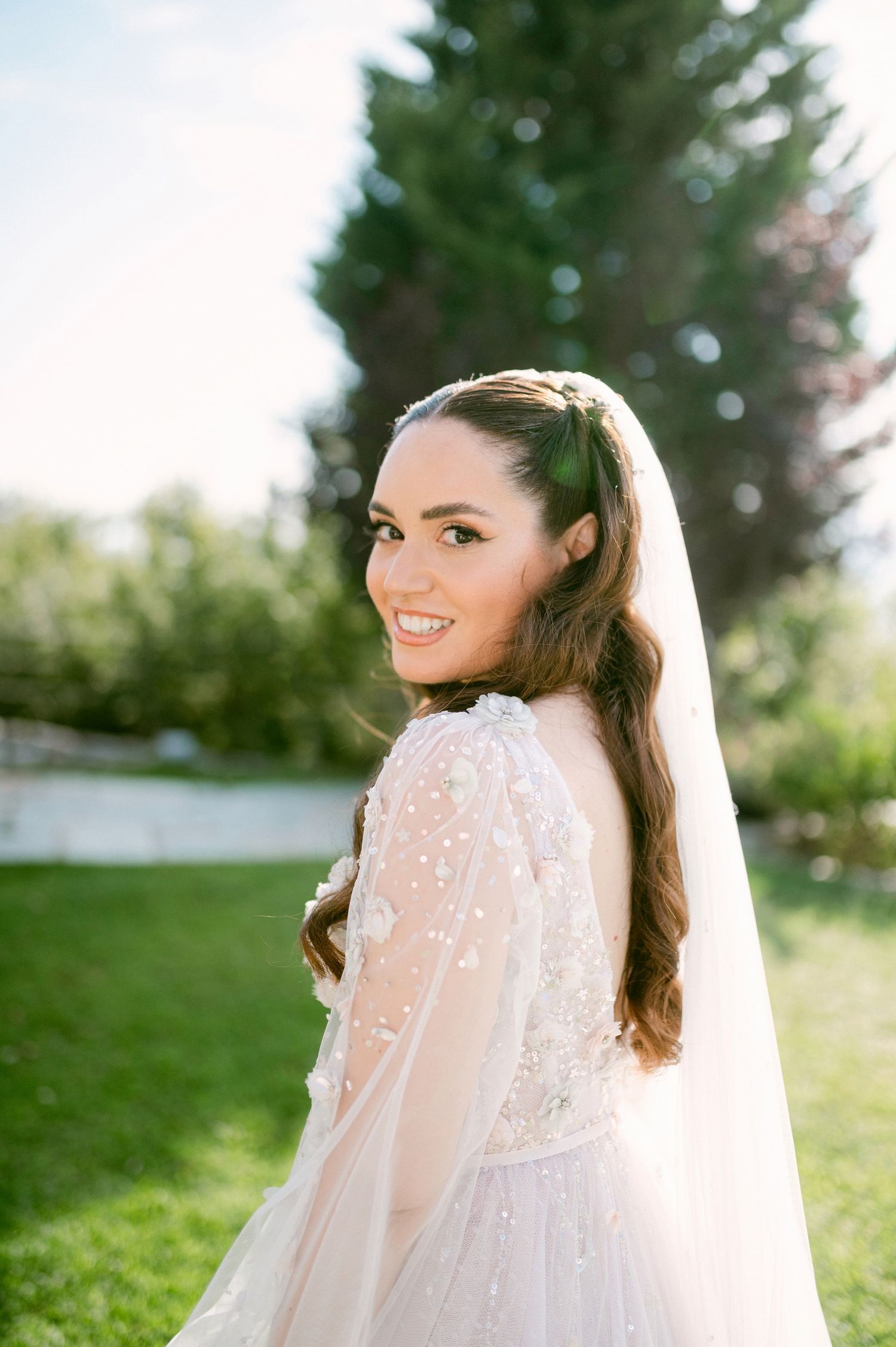 Beautiful bride in a delicate sheer long-sleeved wedding gown with floral details poses outdoors on a sunny day.