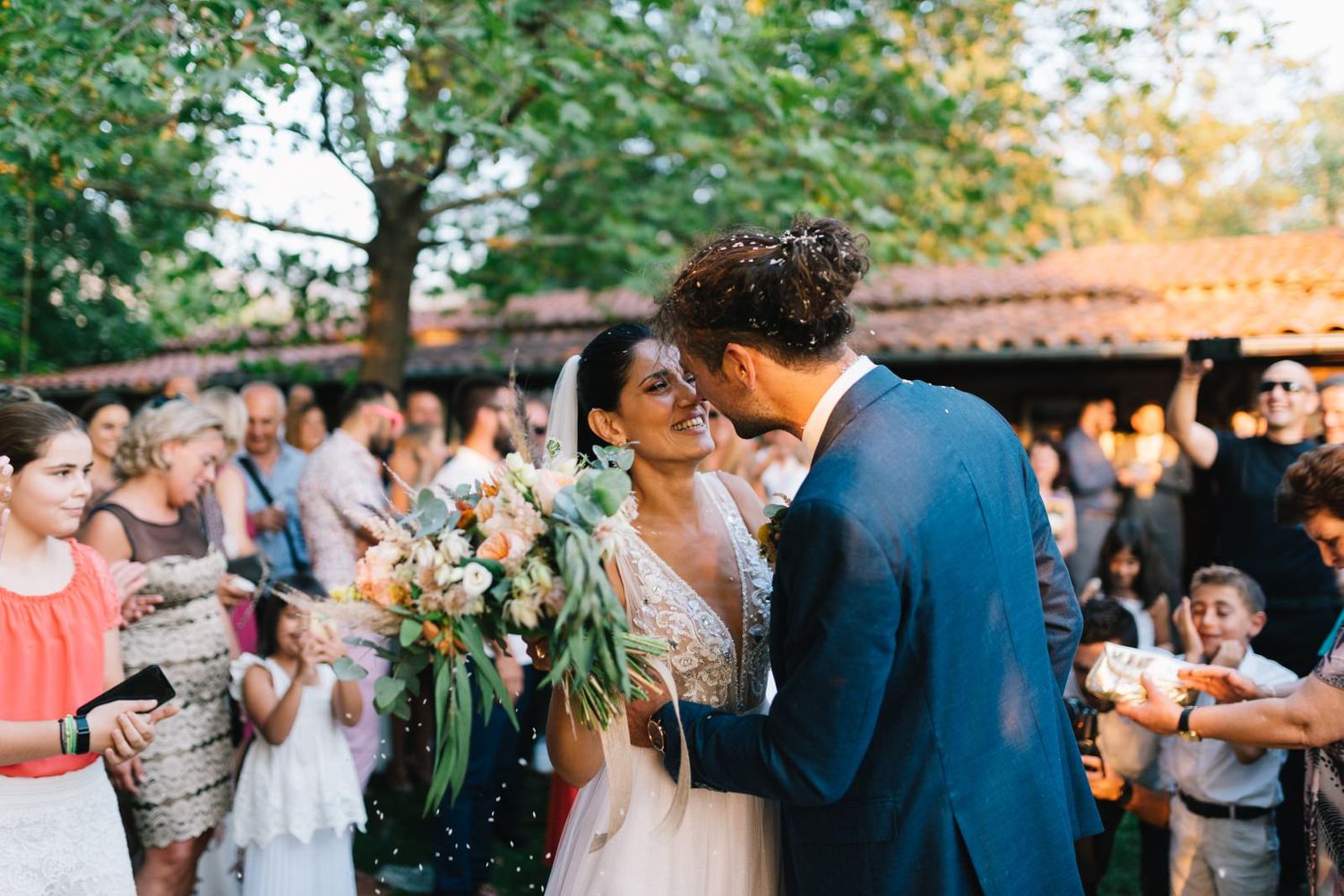Outdoor wedding celebration with guests gathered around newlyweds in a garden setting at sunset.