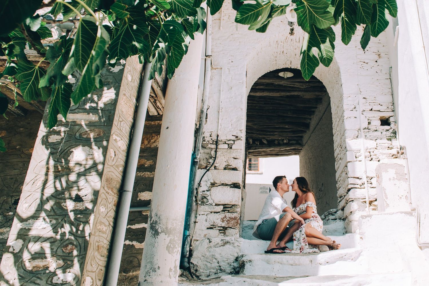 A romantic moment captured in a whitewashed Mediterranean archway with dappled sunlight and shadows.