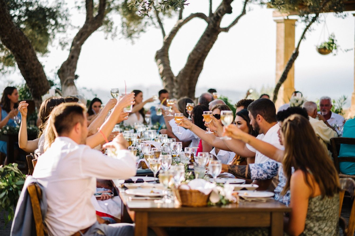 Wedding guests raise their glasses in a toast at an outdoor reception dinner under string lights.