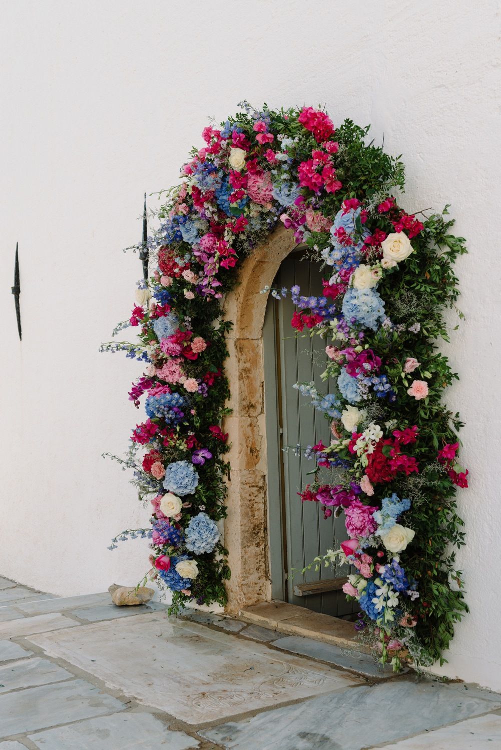 Romantic doorway arch adorned with pink, blue and white flowers against white stone wall.