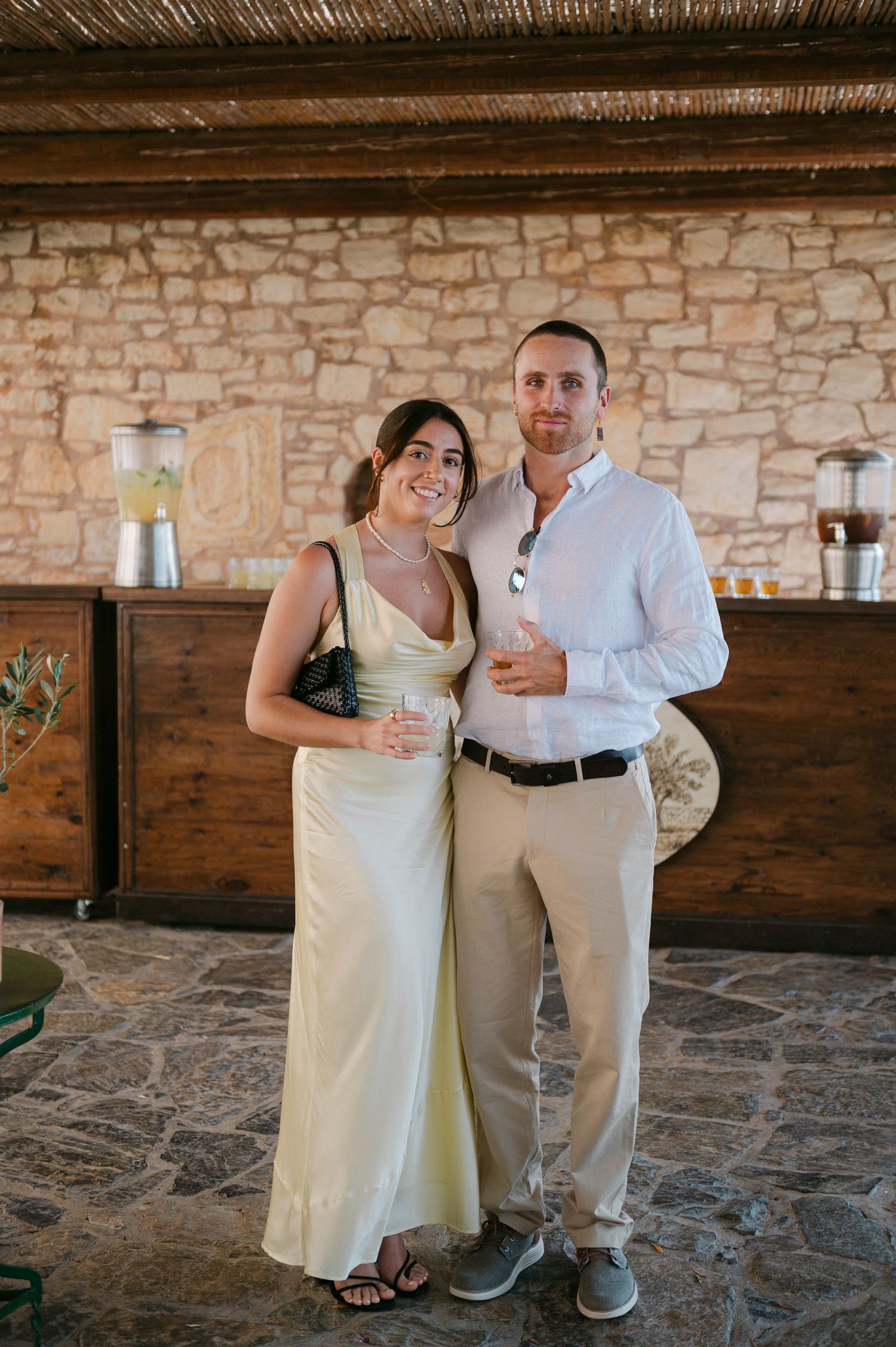 Couple in light colored attire poses together on stone floor against rustic stone wall interior.