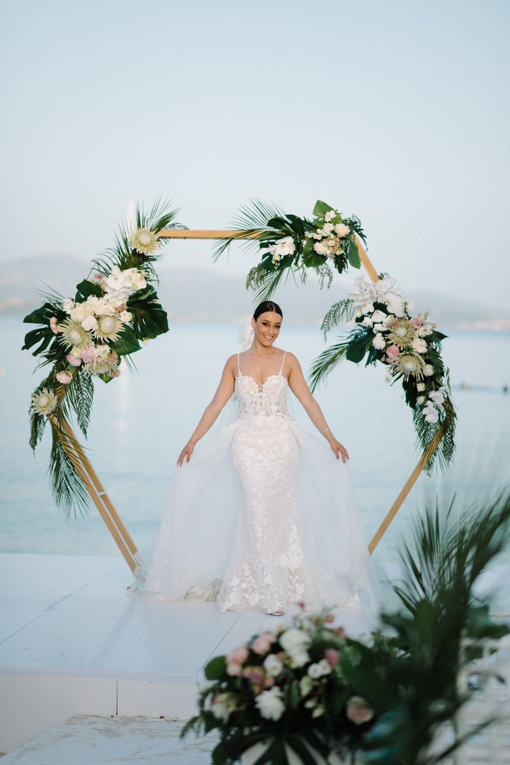 Bride showing off flowing wedding dress under tropical flower arch at seaside venue.