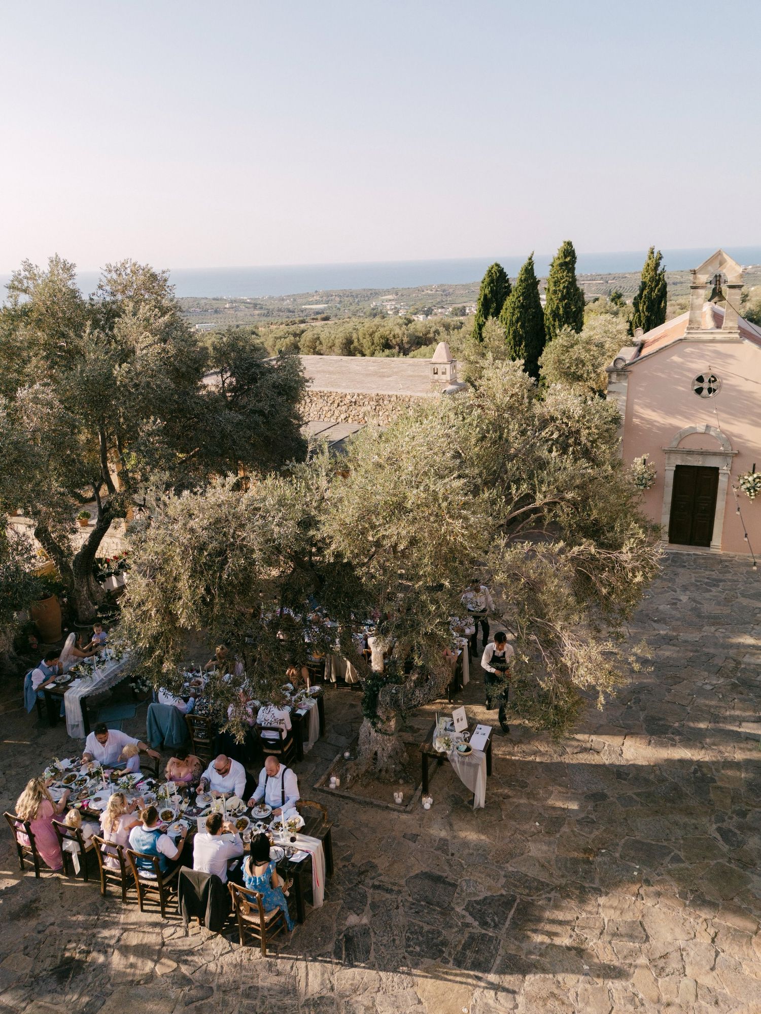 People gathered for an outdoor meal under olive trees beside a rustic stone church overlooking a scenic Mediterranean landscape.