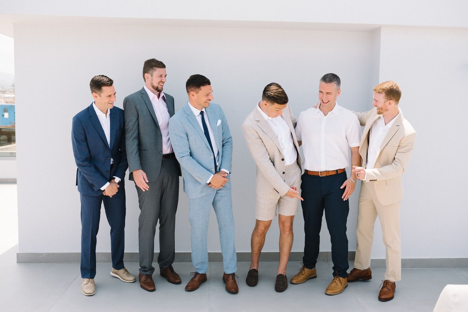 Group of groomsmen in coordinated suits laughing together on a bright terrace.