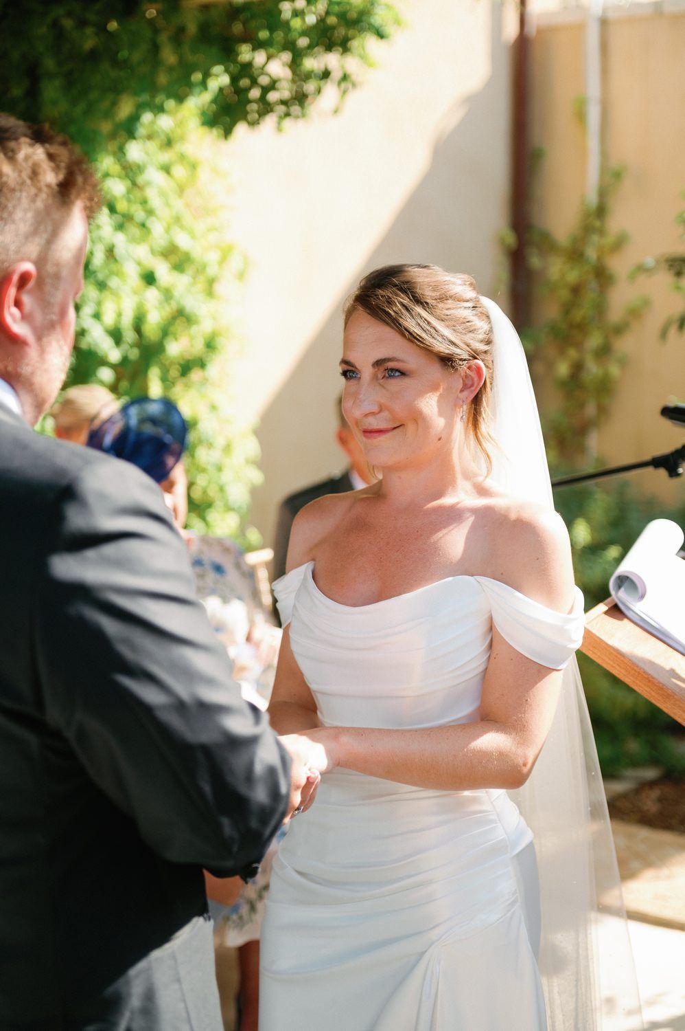 A bride in an off-shoulder white gown exchanges vows during an outdoor ceremony.