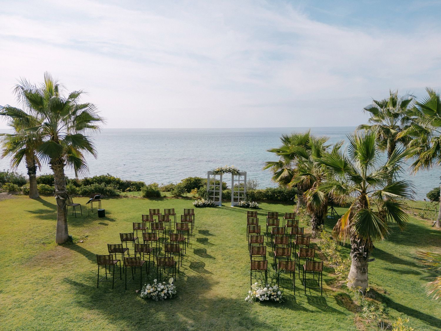 Rows of wedding ceremony chairs arranged on oceanfront lawn with palm trees and gazebo overlooking the sea.