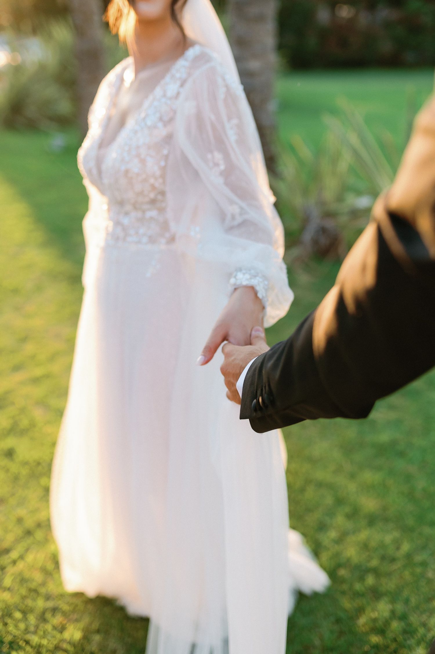 A bride in a flowing white lace wedding dress stands in a sunlit garden setting.