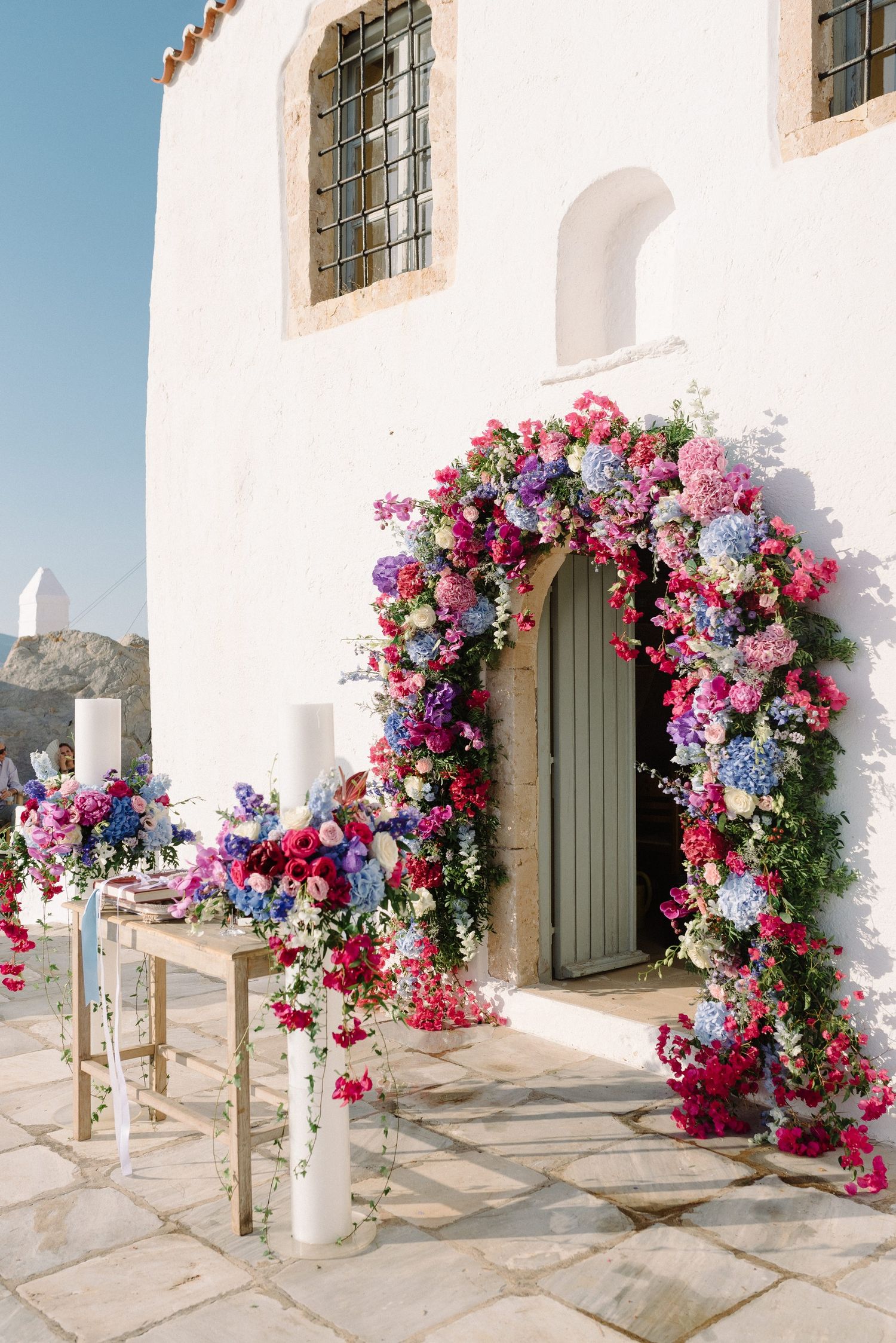 A stunning floral arch with pink and blue blooms adorns a white church doorway in Hydra island.