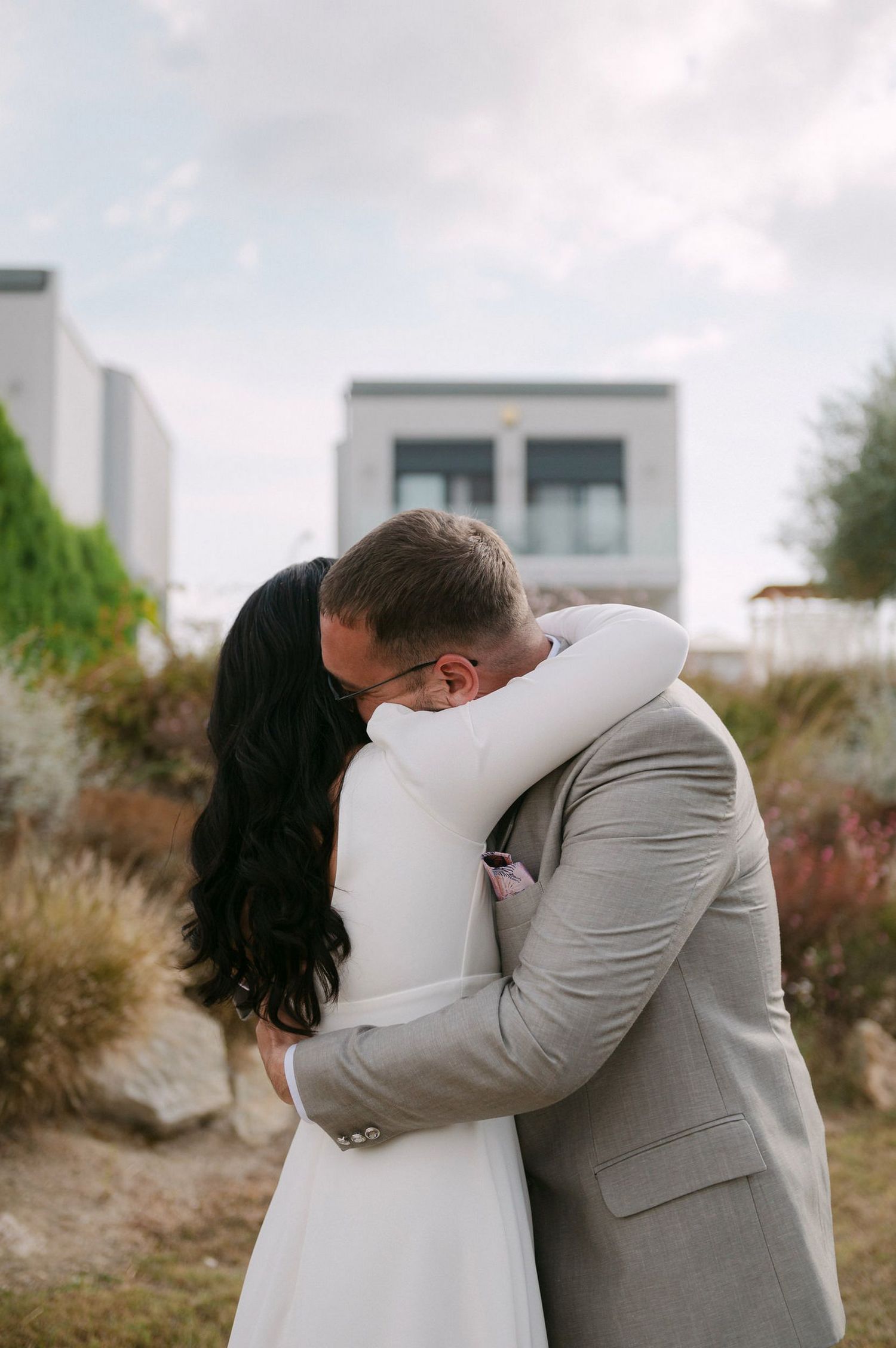A couple shares an intimate embrace outdoors, with the person in a white dress hugging someone wearing a gray suit.