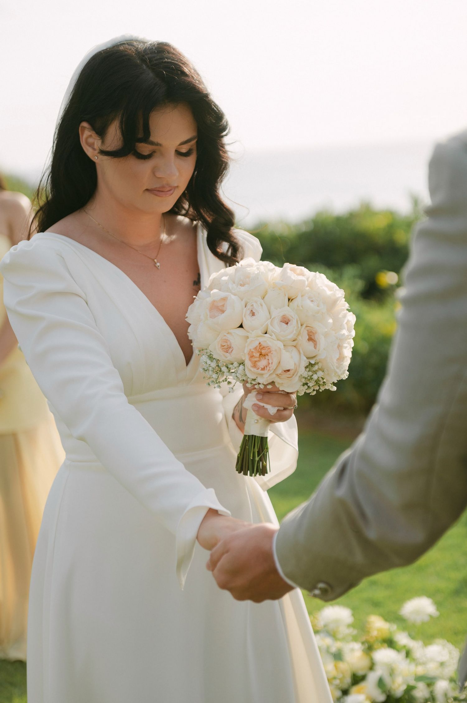 A bride in a long-sleeved white wedding dress holds a beautiful bouquet of cream roses during an outdoor ceremony.