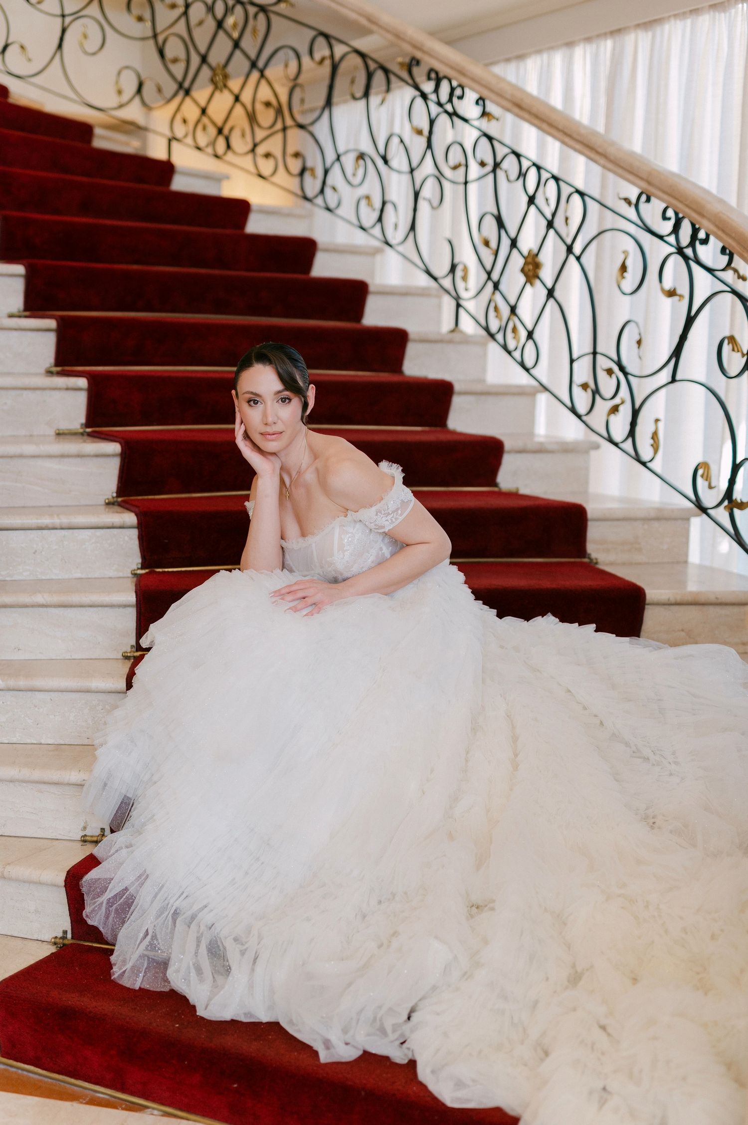 Elegant bride in flowing white gown poses gracefully on grand staircase with red carpet and ornate railing.
