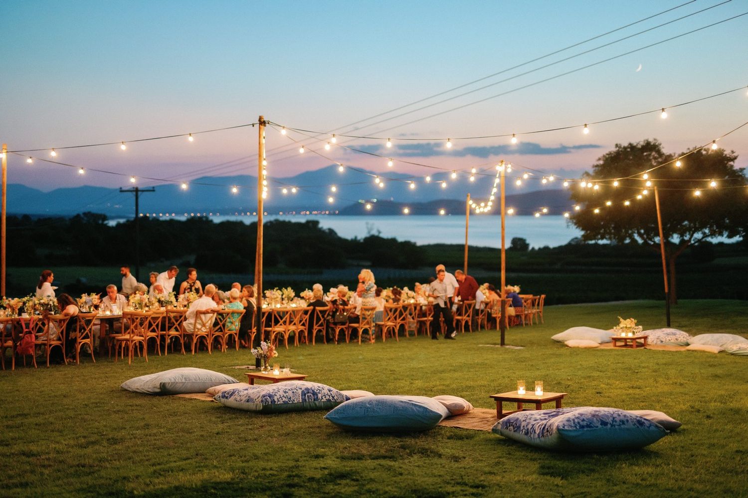 String lights illuminate an outdoor wedding reception setup with long tables and chairs in a rustic field setting at dusk.