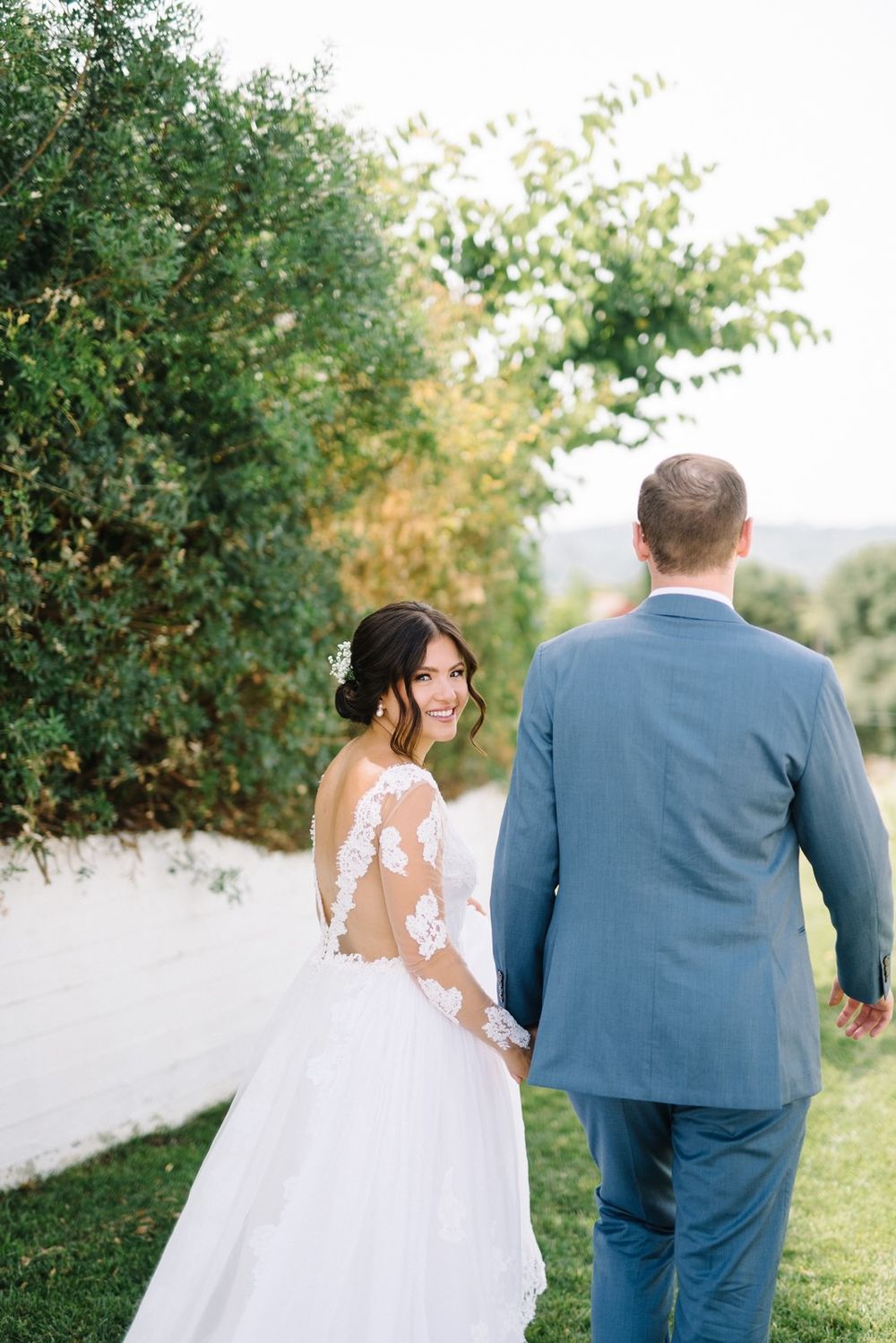 Couple in wedding attire walking through a lush garden pathway with greenery and white flowers.