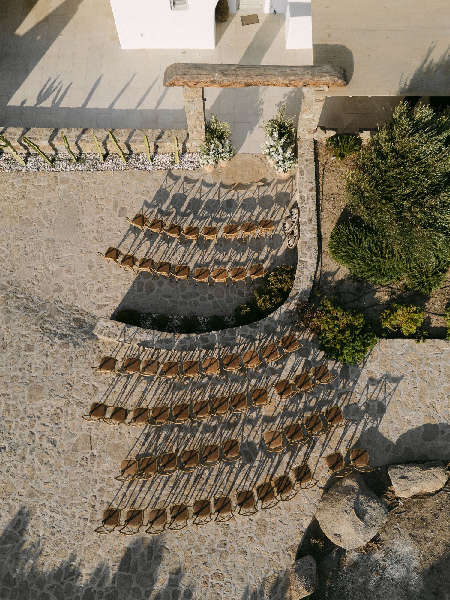 Aerial view of empty wooden beach chairs casting dramatic shadows in a curved pattern on sandy ground.