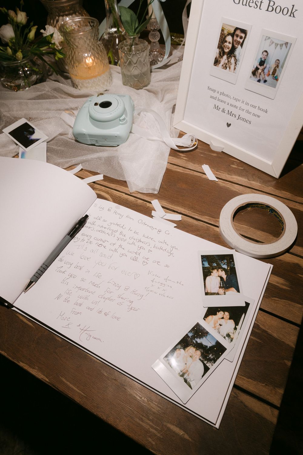 Wedding guest book setup with Instax camera, handwritten messages, and photos on rustic wooden table with candles.