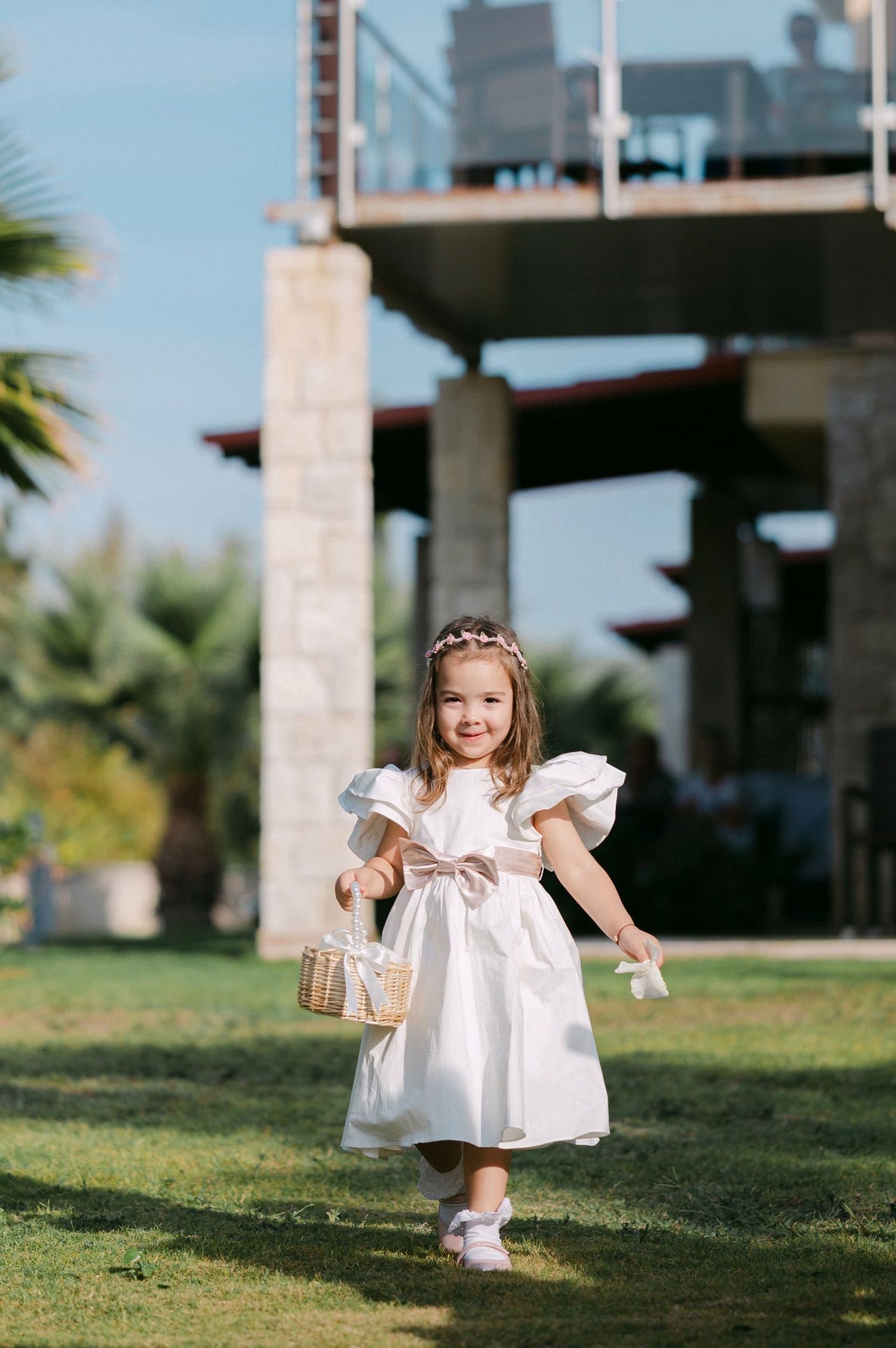 A small child in a white dress plays on a grassy lawn outside a modern building on a sunny day.