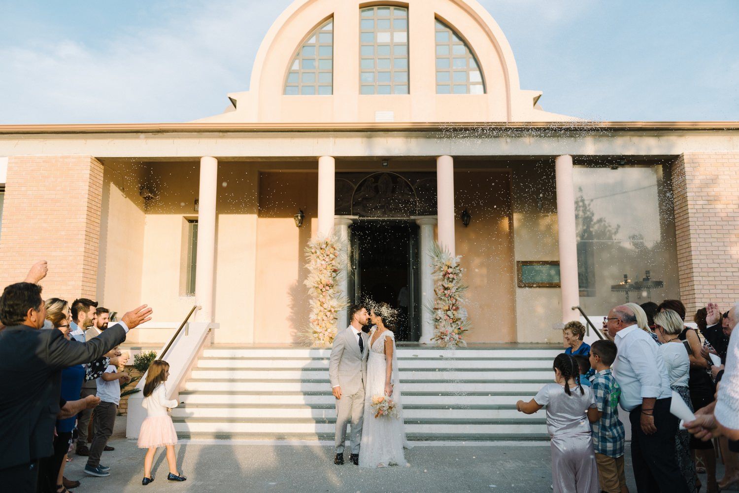 Wedding guests gather outside a church with white columns as confetti fills the air.