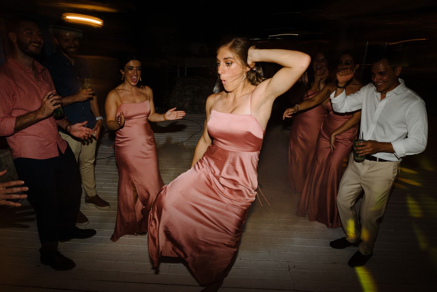 Wedding party members dancing in pink dresses at a dimly lit reception.