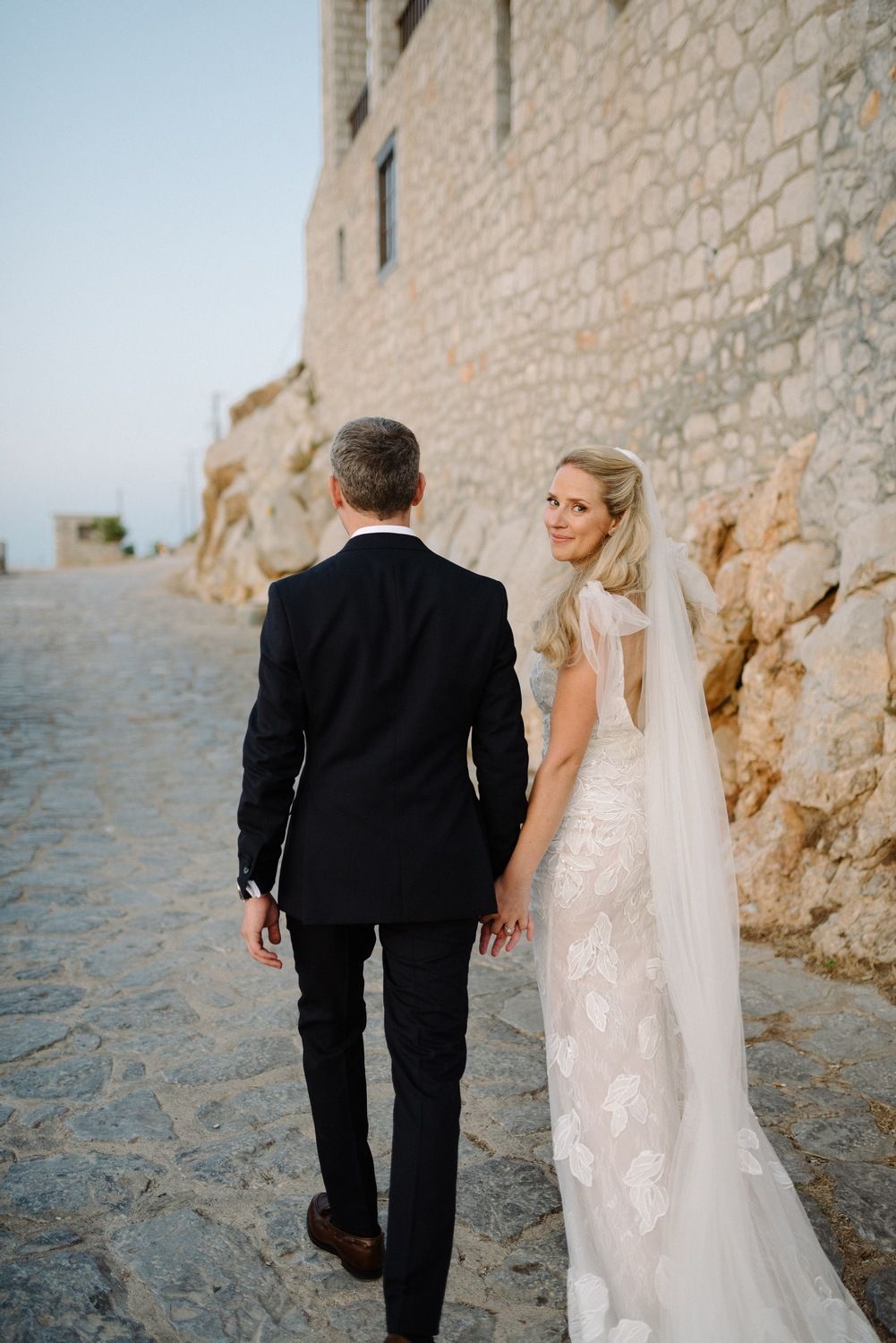 Couple in wedding attire walking along scenic coastal path beside stone building.