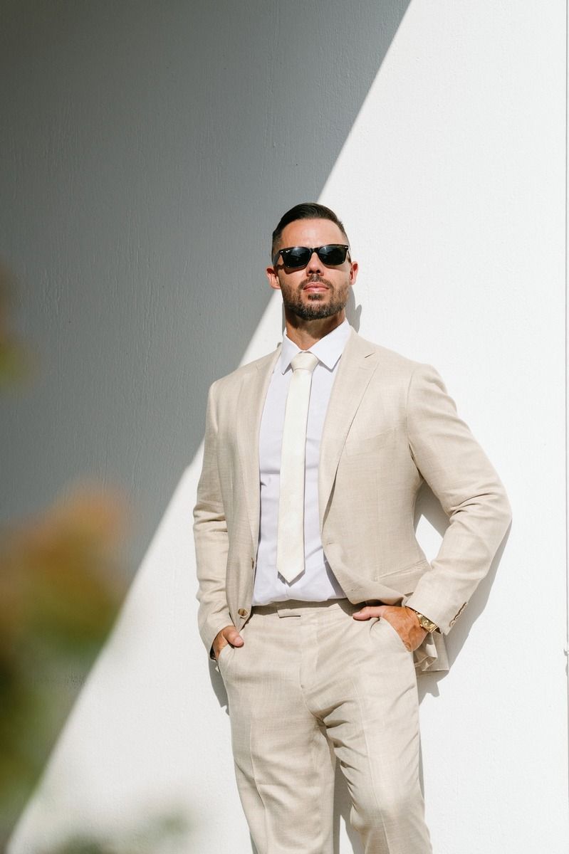 Well-dressed man in cream colored suit poses confidently in dramatic lighting.