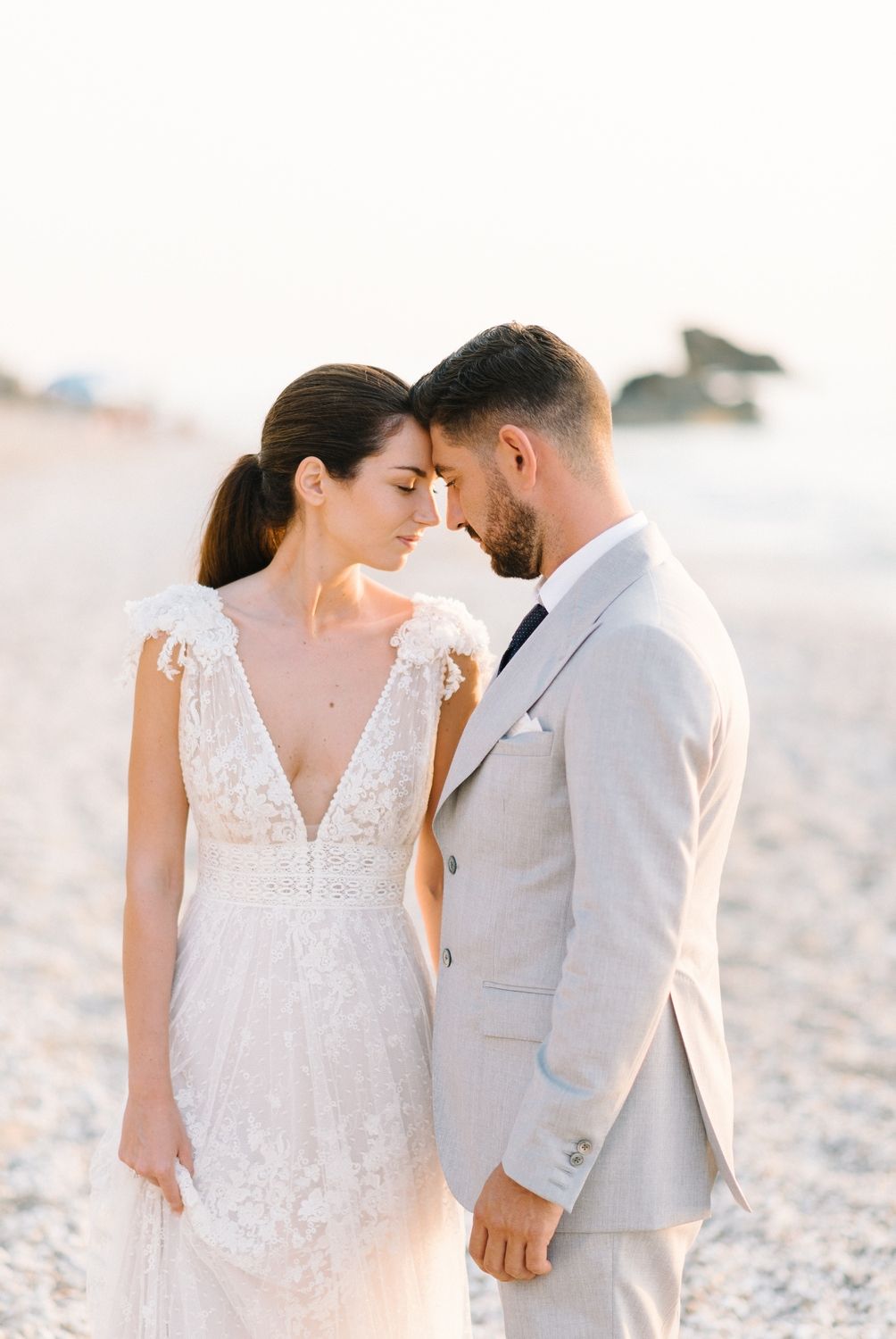 A tender beach wedding moment shows a couple in elegant white and grey attire sharing an intimate moment.