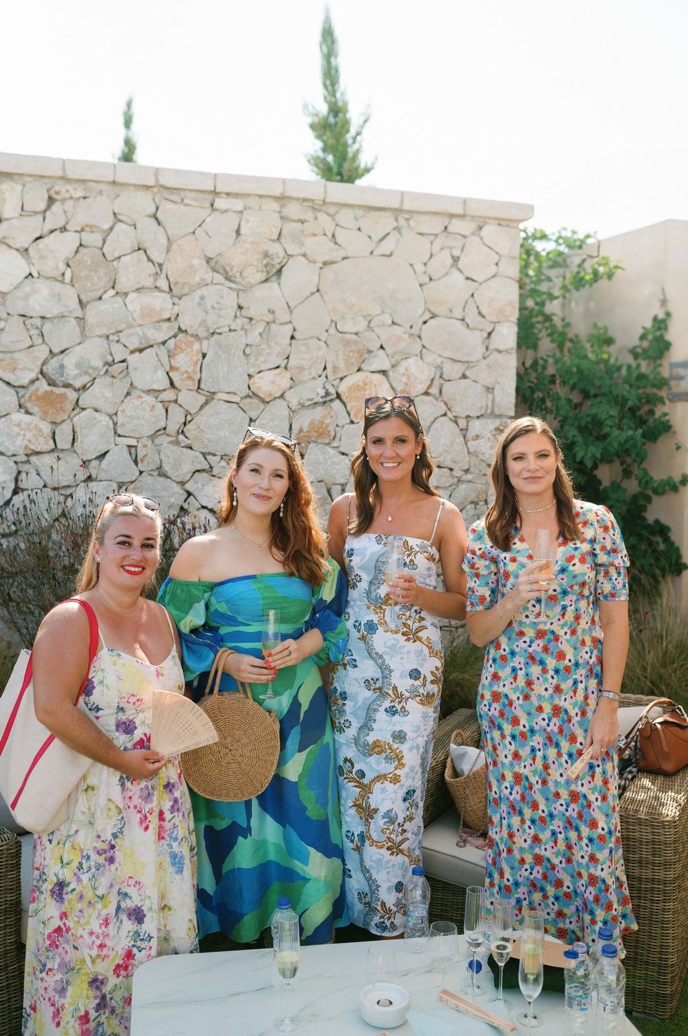 A group of wedding guests in colorful floral dresses pose together against a stone wall.