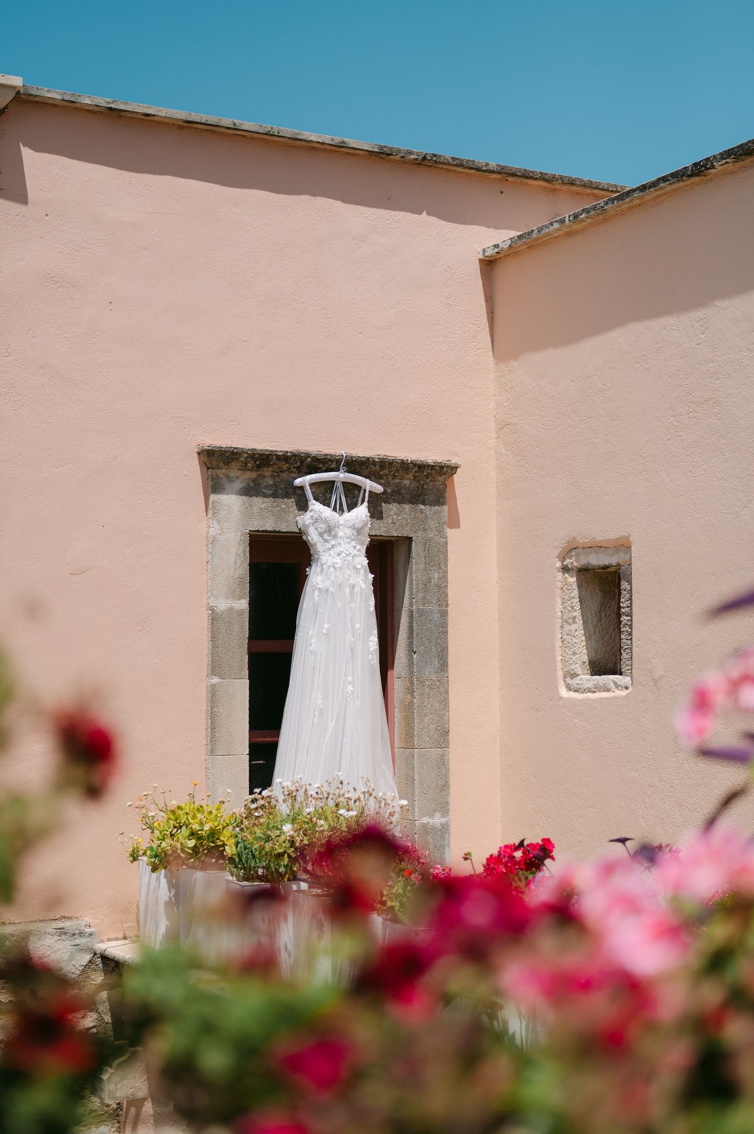 A white wedding dress hangs in a stone doorway of a pink stucco building with pink flowers in the foreground.