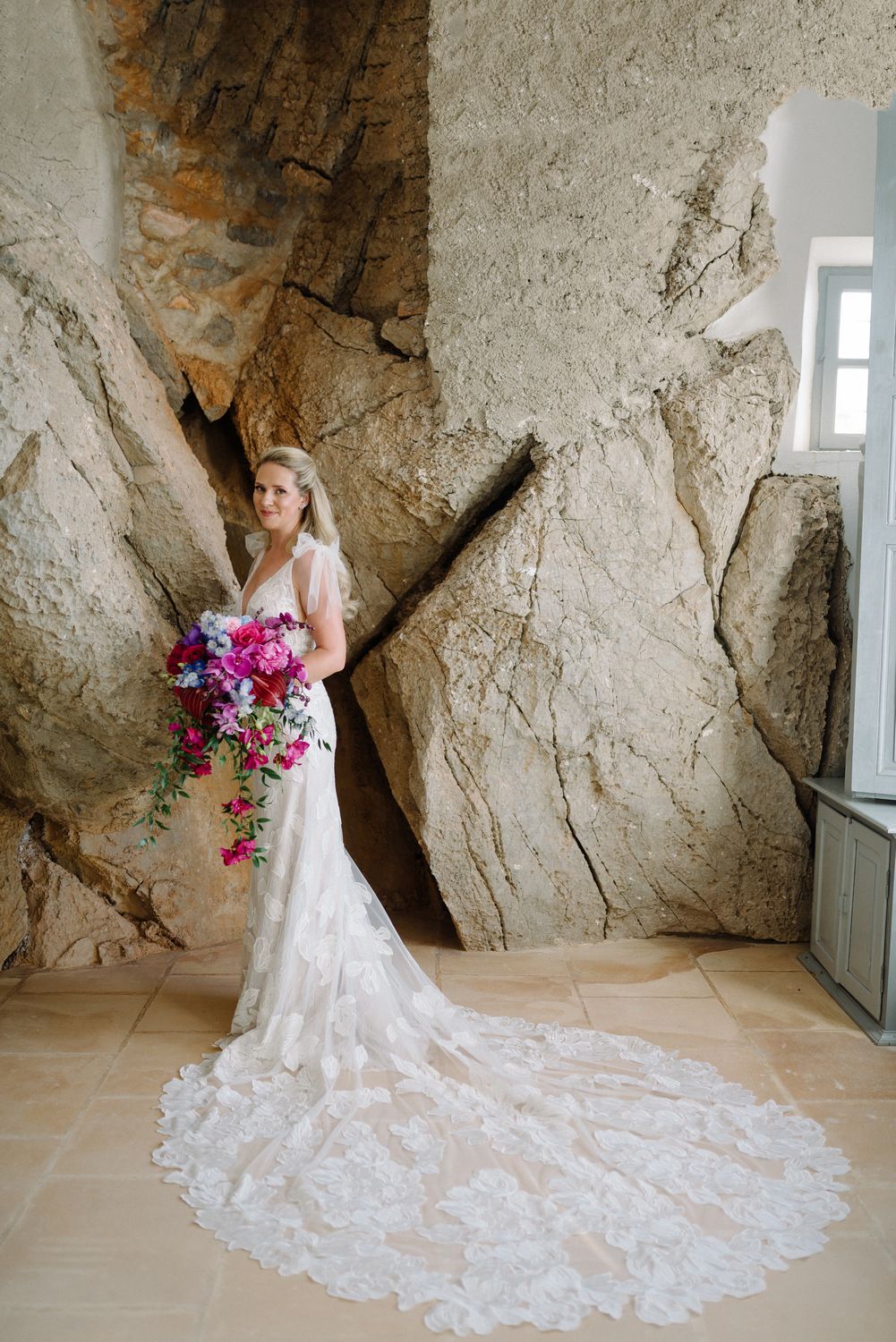 A bride in a long lace train gown holds vibrant pink bouquet against textured stone wall backdrop.