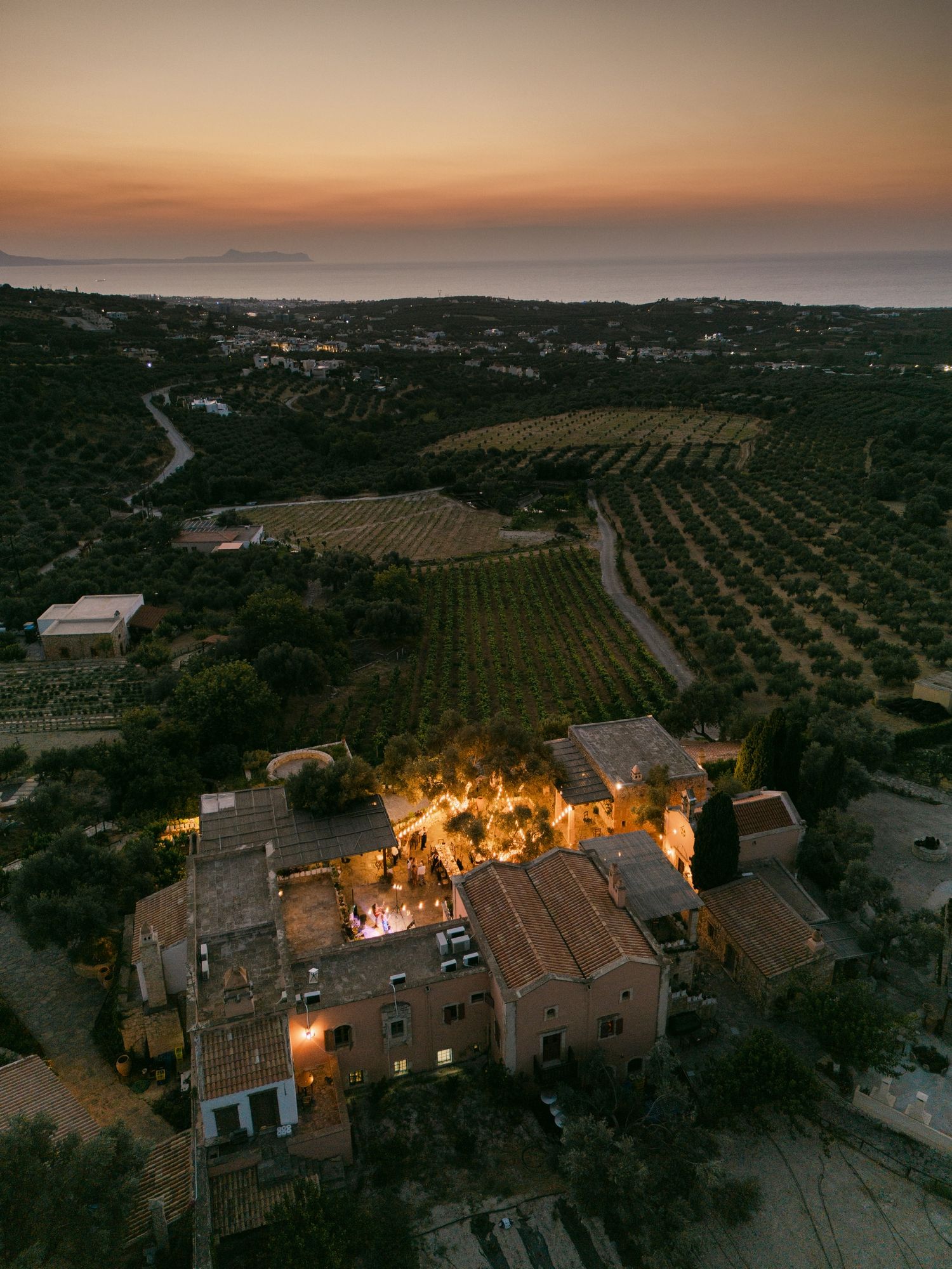 Aerial view of a Mediterranean villa with warm glowing lights at sunset surrounded by terraced olive groves and vineyards.